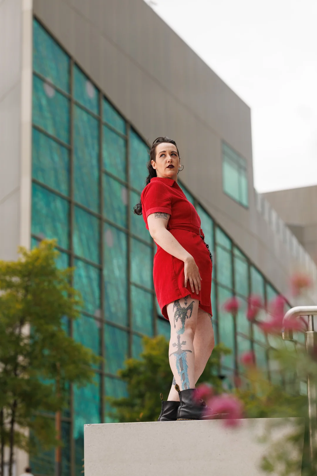 A woman in a red dress with leg tattoos posing against a modern glass building.