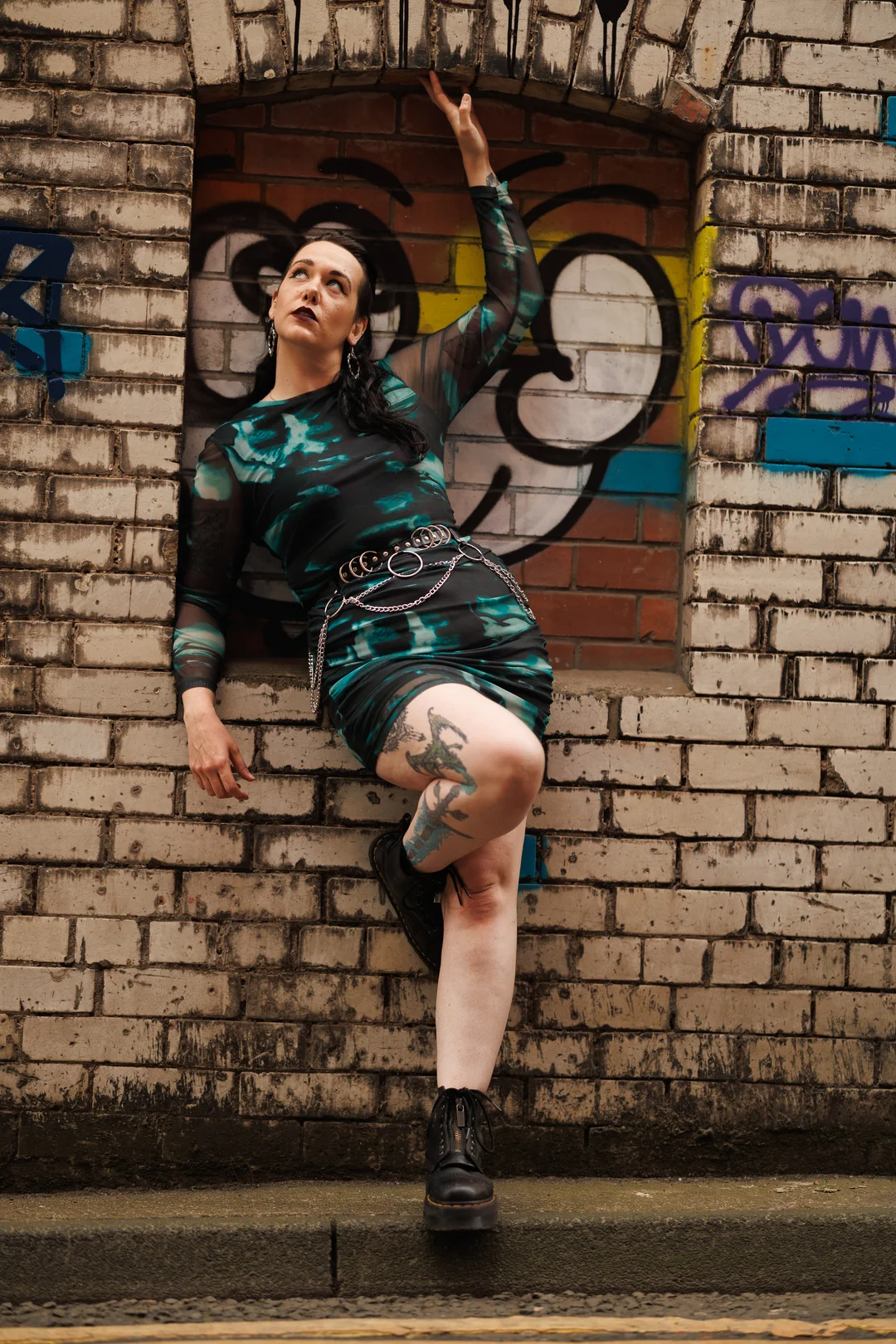 Woman with tattoos posing in a teal and black dress against a graffiti brick wall.