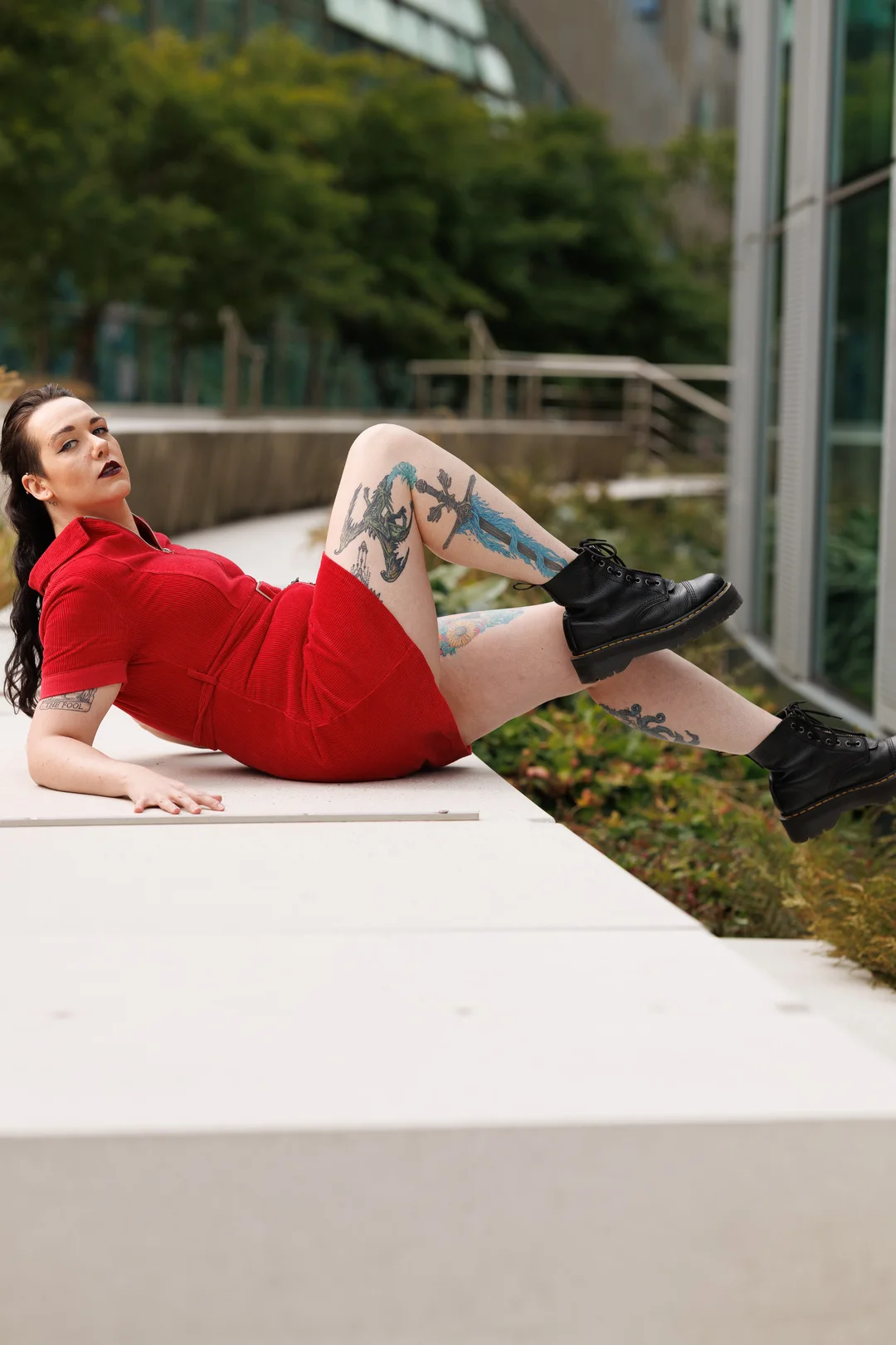 Woman with tattoos in a red dress and black boots reclining on a white ledge in an outdoor urban setting.