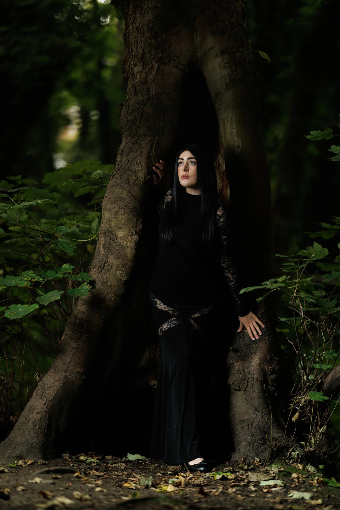 A woman in a long black dress standing between the roots of a large tree in a moody, dark forest.