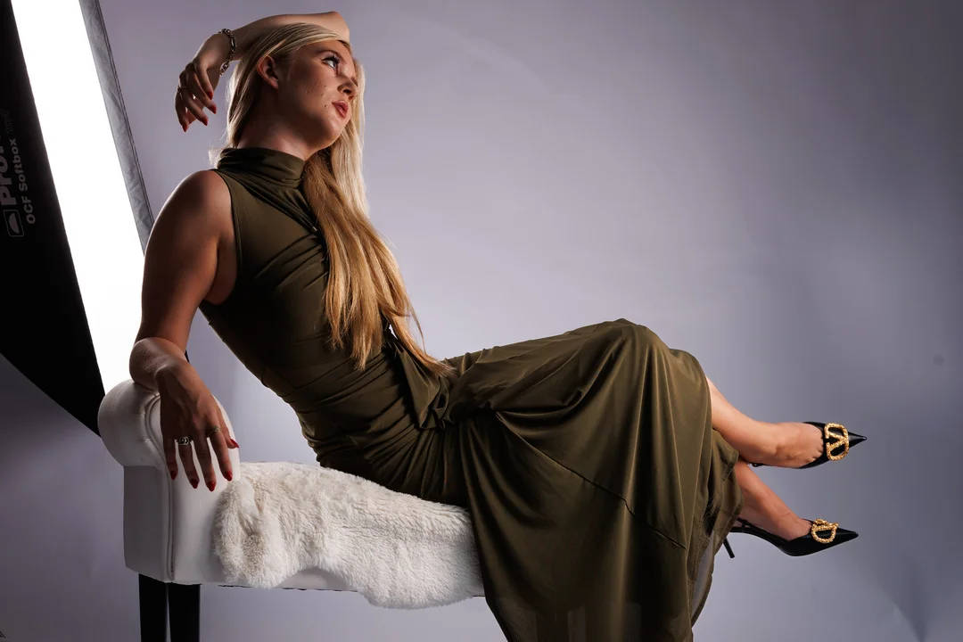 Blonde woman in an olive green dress posing on a white faux-fur chair in a studio setting.
