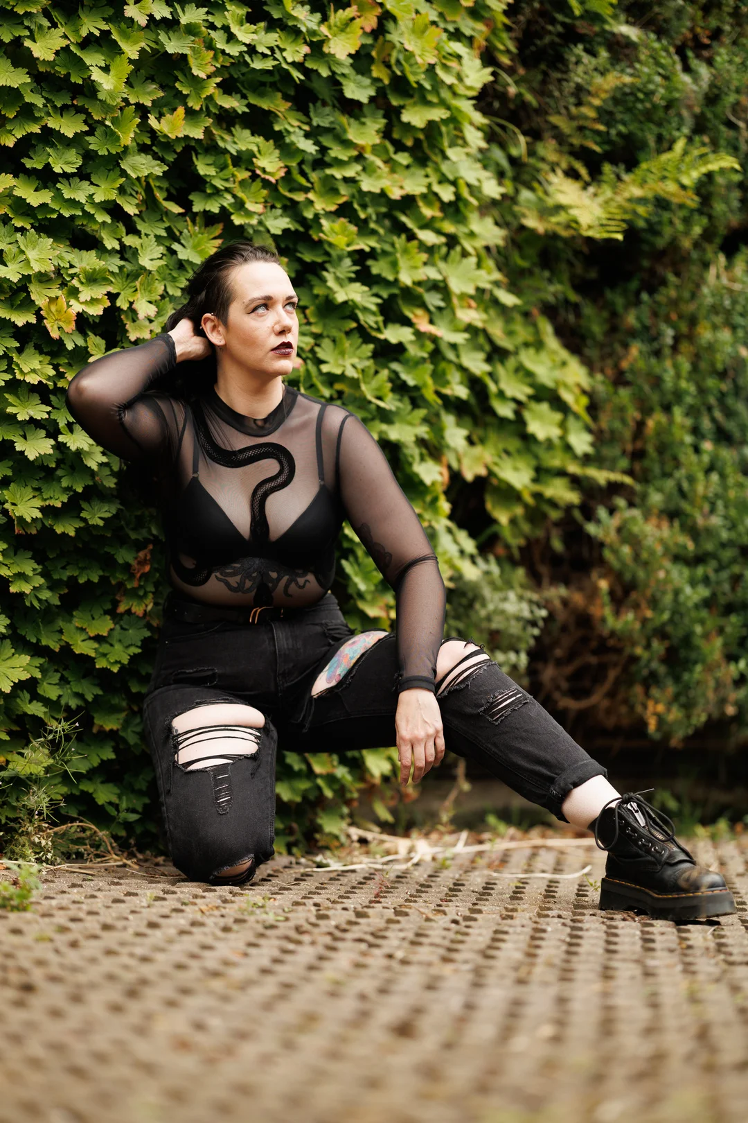 Woman in a black sheer snake-print top and ripped black jeans posing against a lush green hedge.