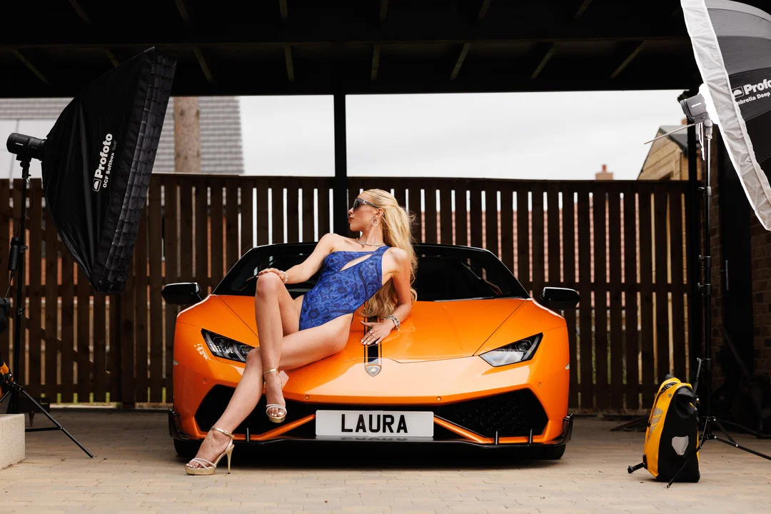 Blonde woman in a blue swimsuit posing on an orange Lamborghini with studio lighting.