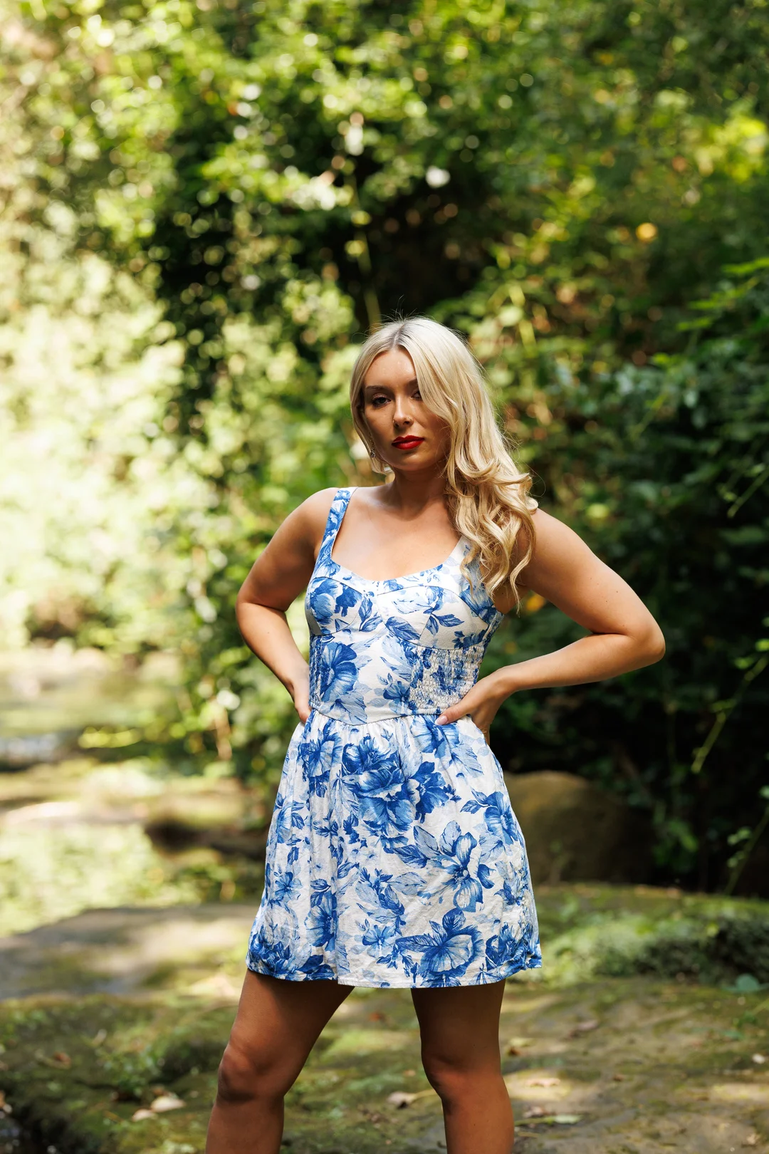 A blonde woman in a blue and white floral dress posing in a lush green forest setting.