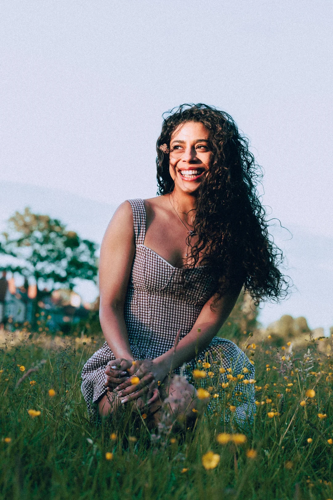 Smiling woman with curly hair in a gingham dress crouching in a field of yellow wildflowers.