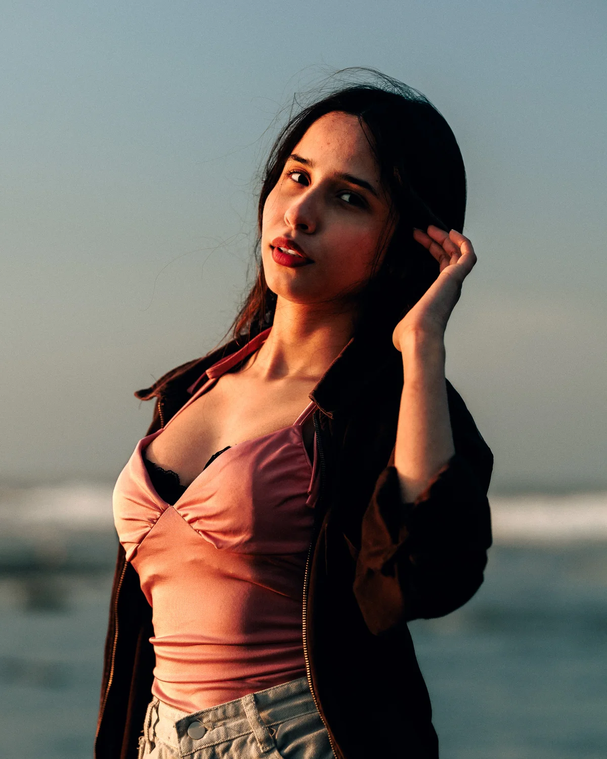 A young woman with dark hair and red lipstick posing outdoors during golden hour, wearing a pink top and dark jacket.