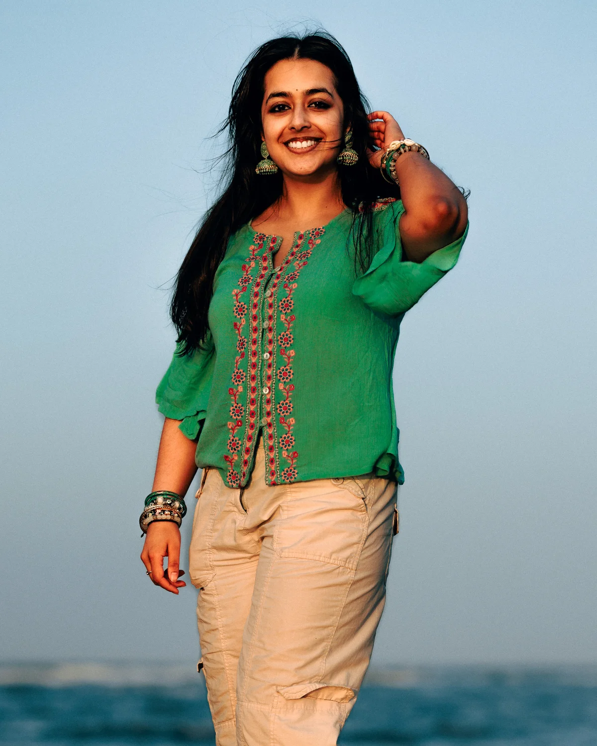 A smiling woman in a green embroidered top and beige cargo pants standing on a beach under a clear sky.