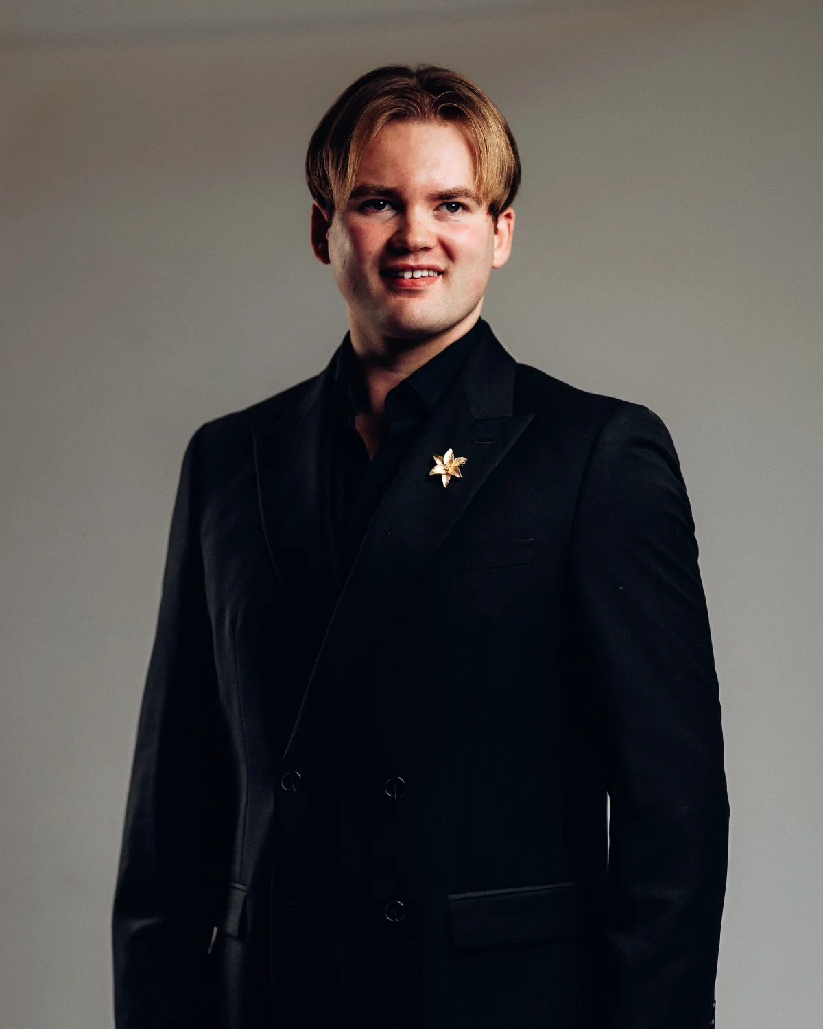 A man with blond hair in a black double-breasted suit and gold lapel pin posing against a neutral grey background.