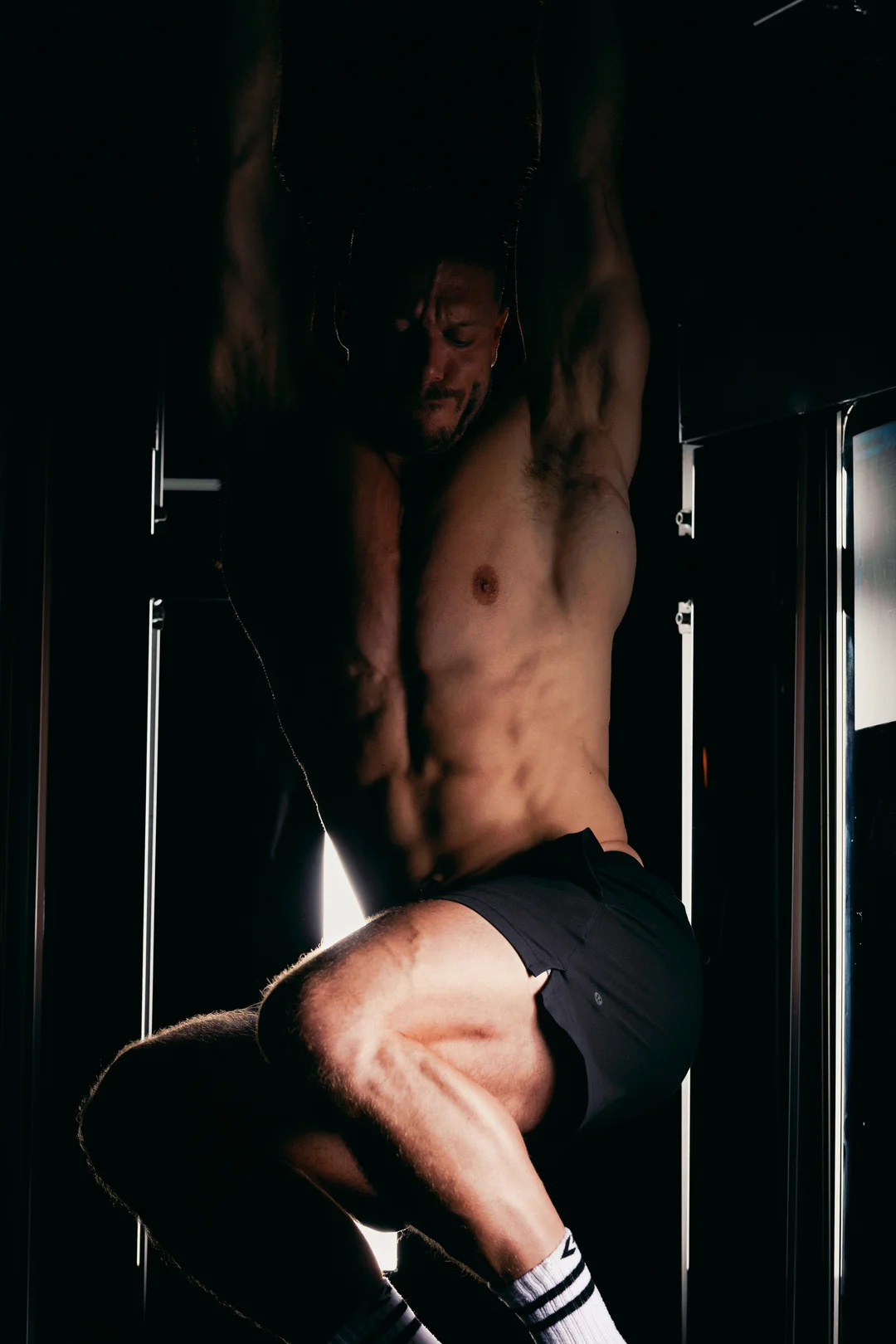 Dramatic low-key photograph of a shirtless muscular man in a gym, emphasizing muscle definition.