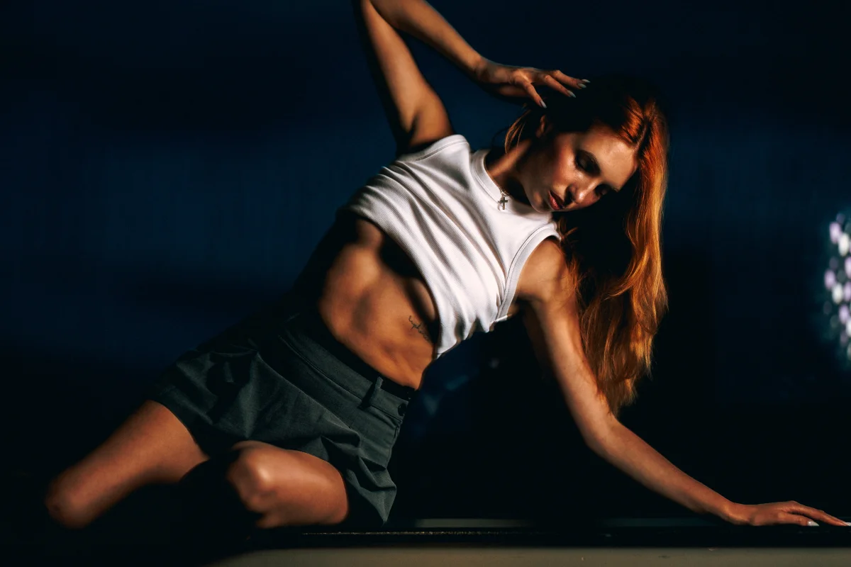 A woman in a white crop top and grey shorts posing dynamically against a dark background with dramatic lighting.