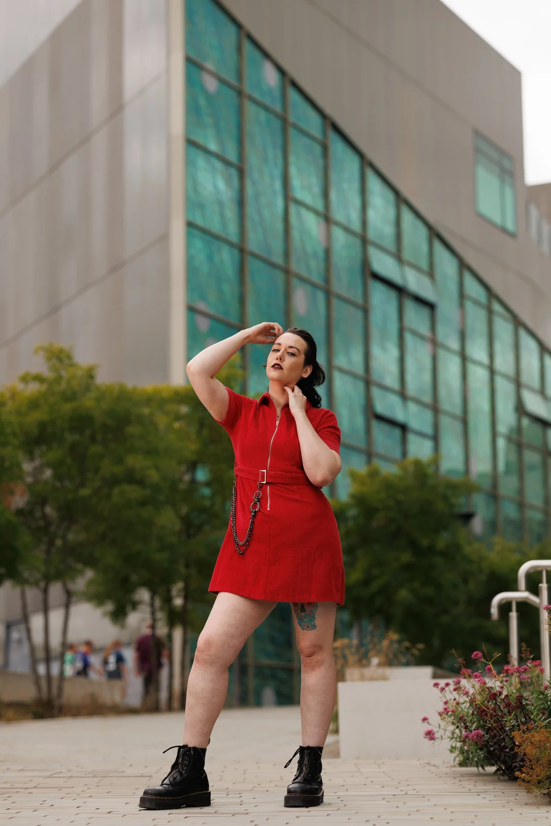 A woman in a short red dress and black boots posing confidently before a modern glass building.
