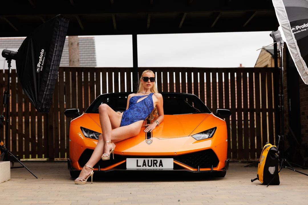 Woman in a blue swimsuit and sunglasses posing on the hood of an orange Lamborghini.