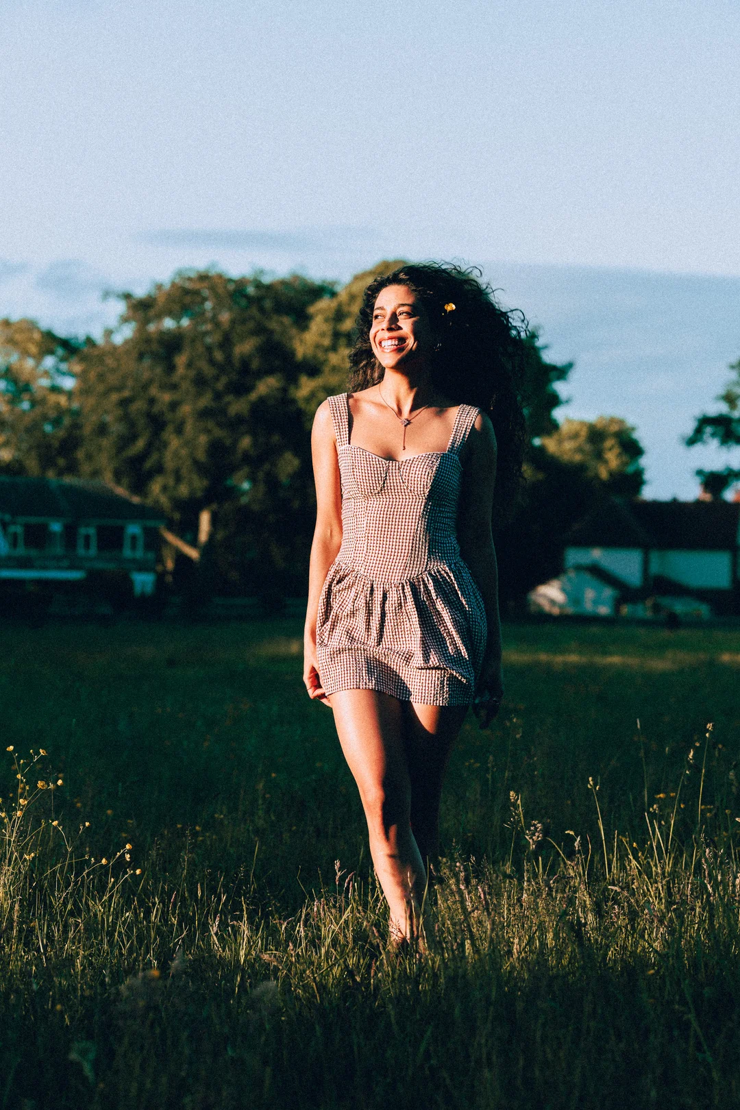 Smiling woman with curly hair in a red gingham dress standing in a sunny grassy field.