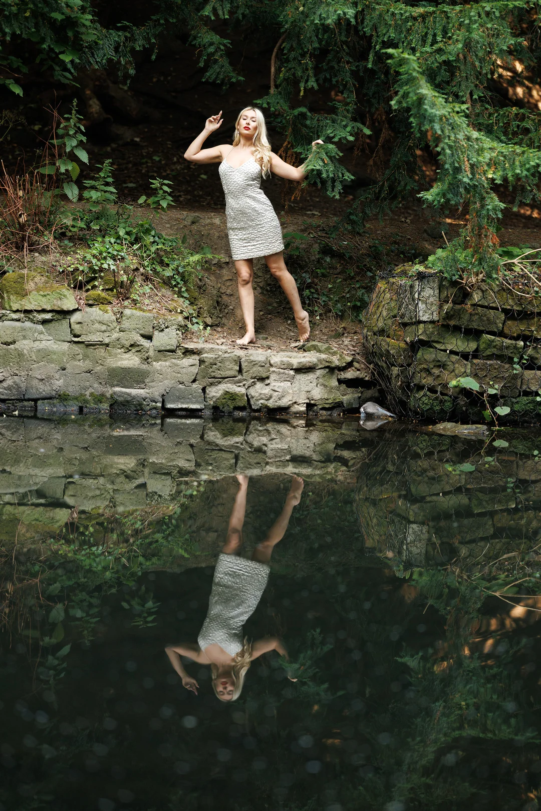 Woman in a white sequined dress posing on a stone ledge with a mirror-like reflection in a pond.