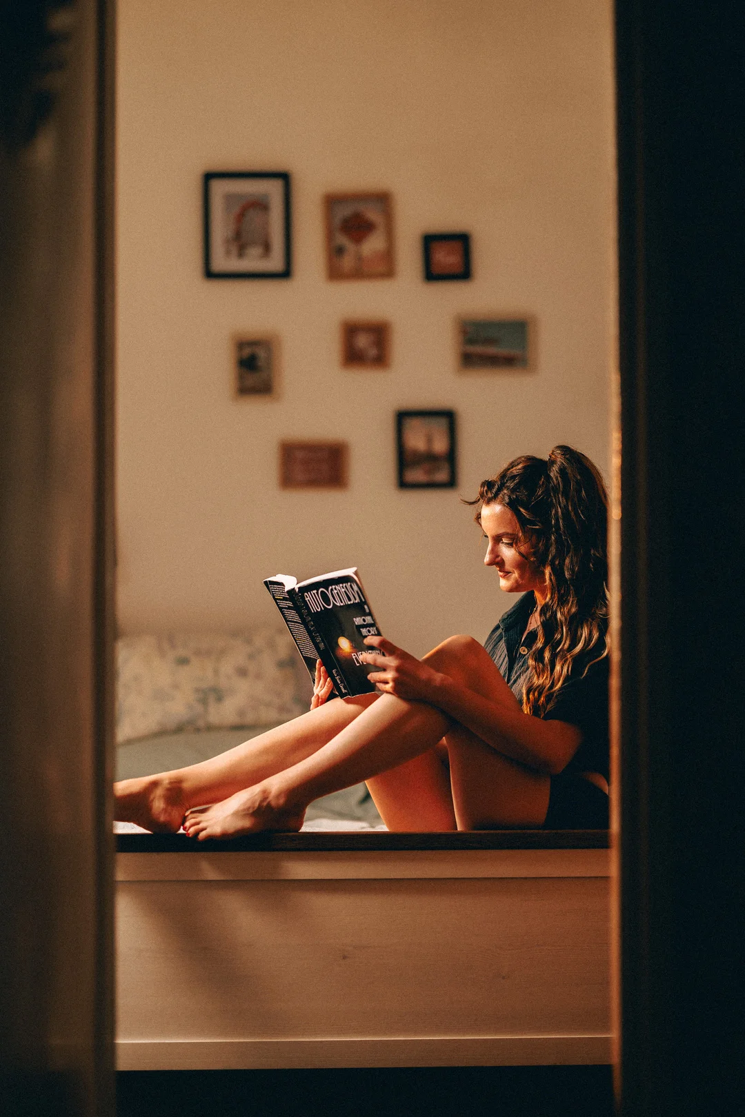 A woman with long curly hair sitting on a bed reading a book in a moody, dimly lit room.