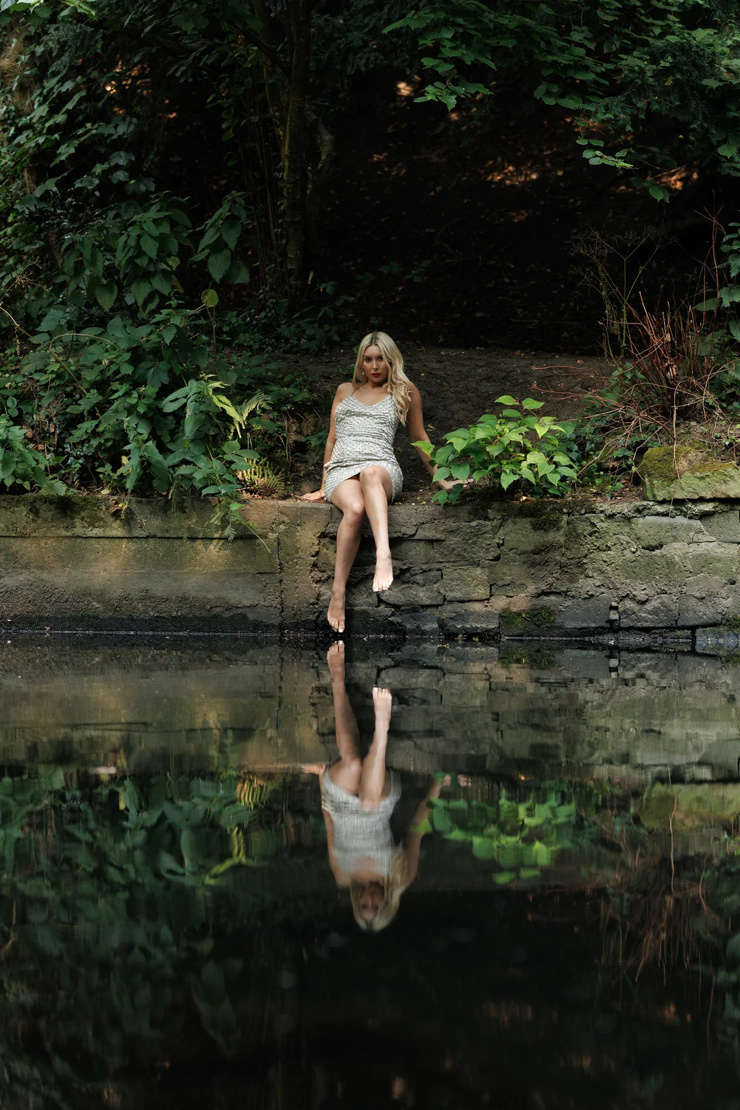 Blonde woman in a silver dress sitting on a stone ledge by a reflective pond in a wooded area.