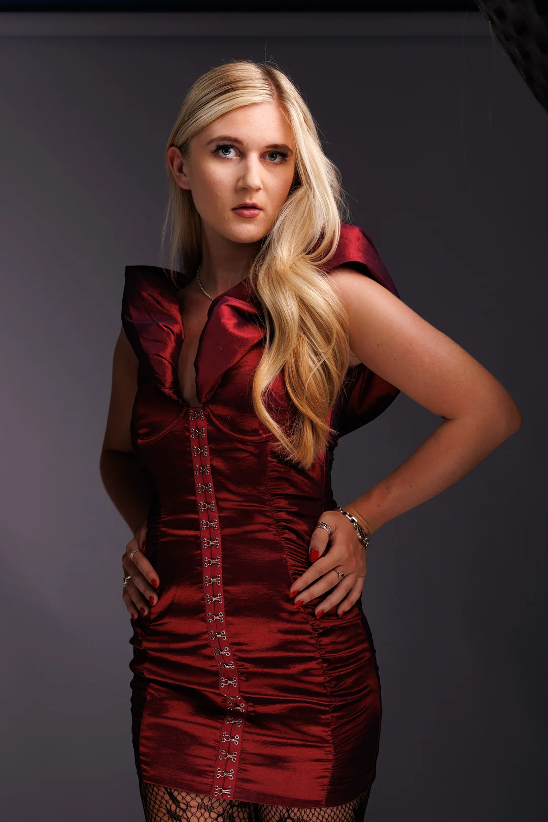 Blonde woman in a structured red satin dress posing against a grey studio background.