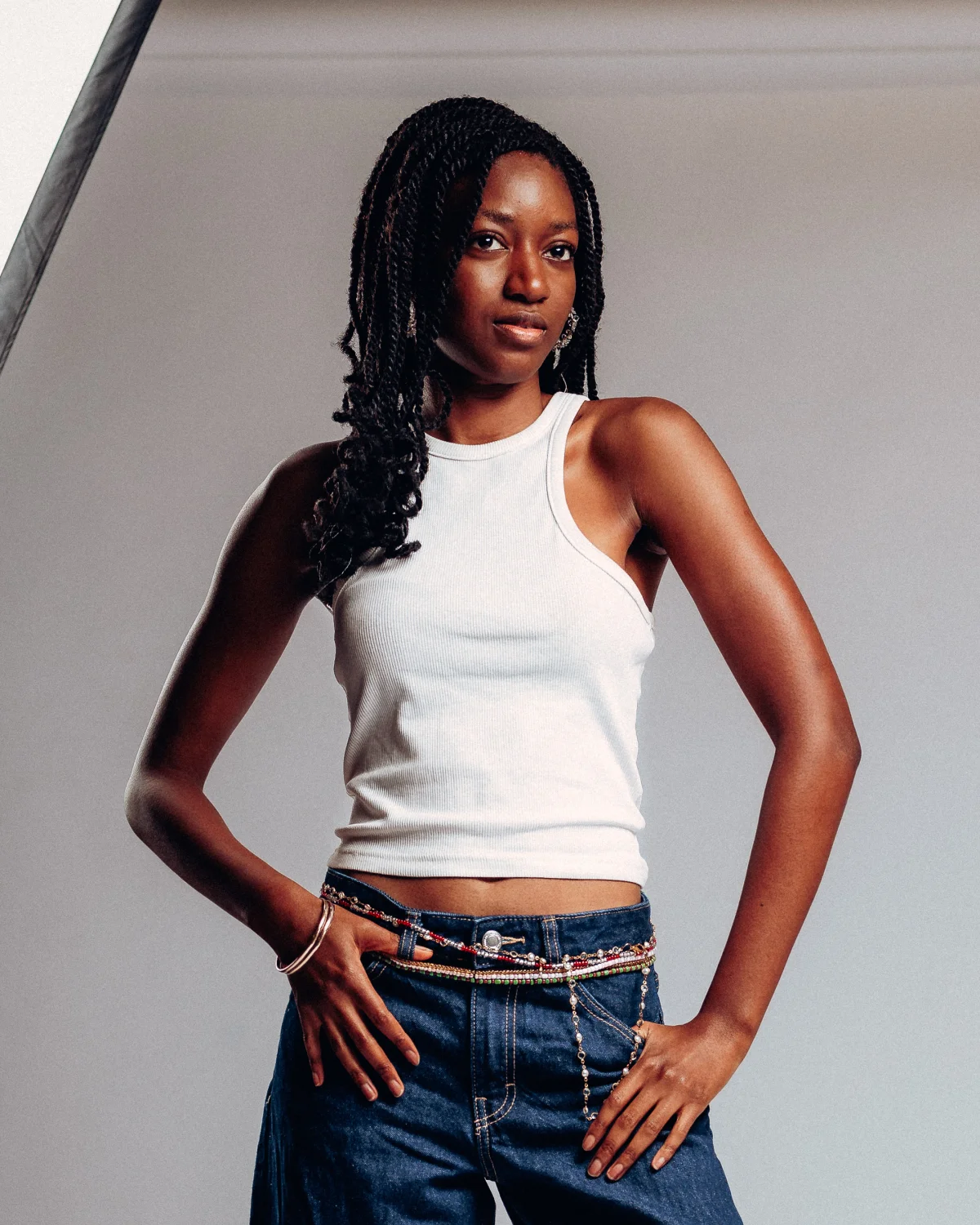 Studio portrait of a Black woman with long braids wearing a white tank top and blue jeans with a beaded belt.