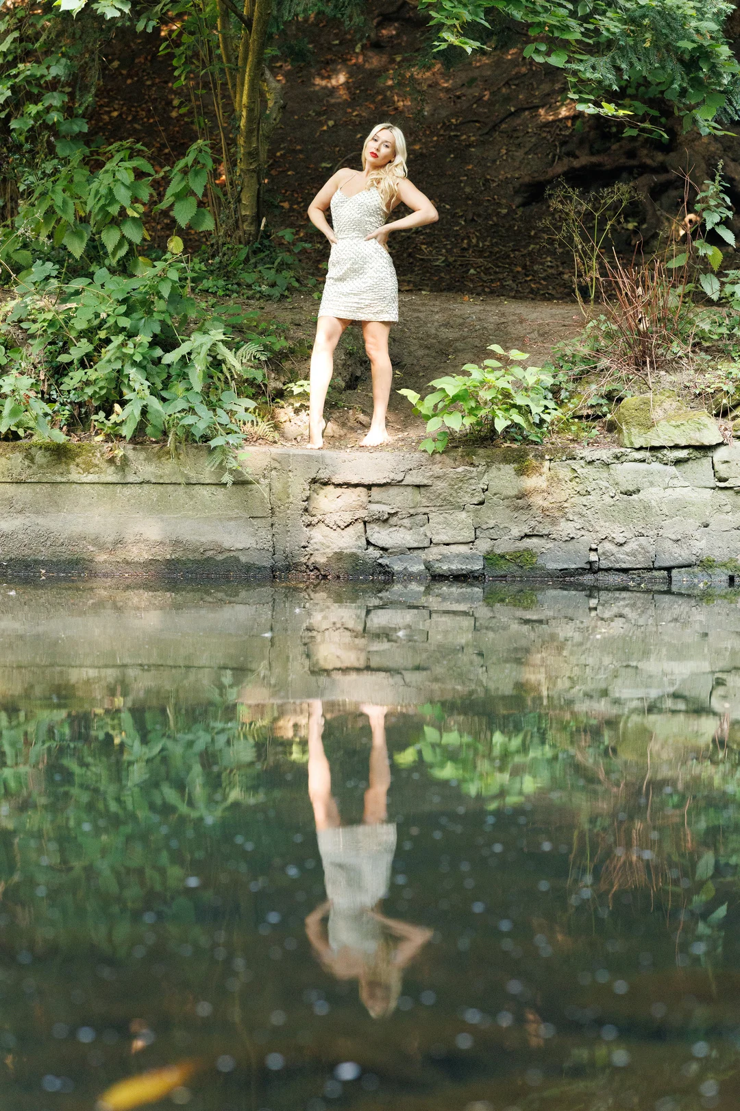 Blonde woman in a sparkly white mini dress posing by a pond, with her reflection in the water.
