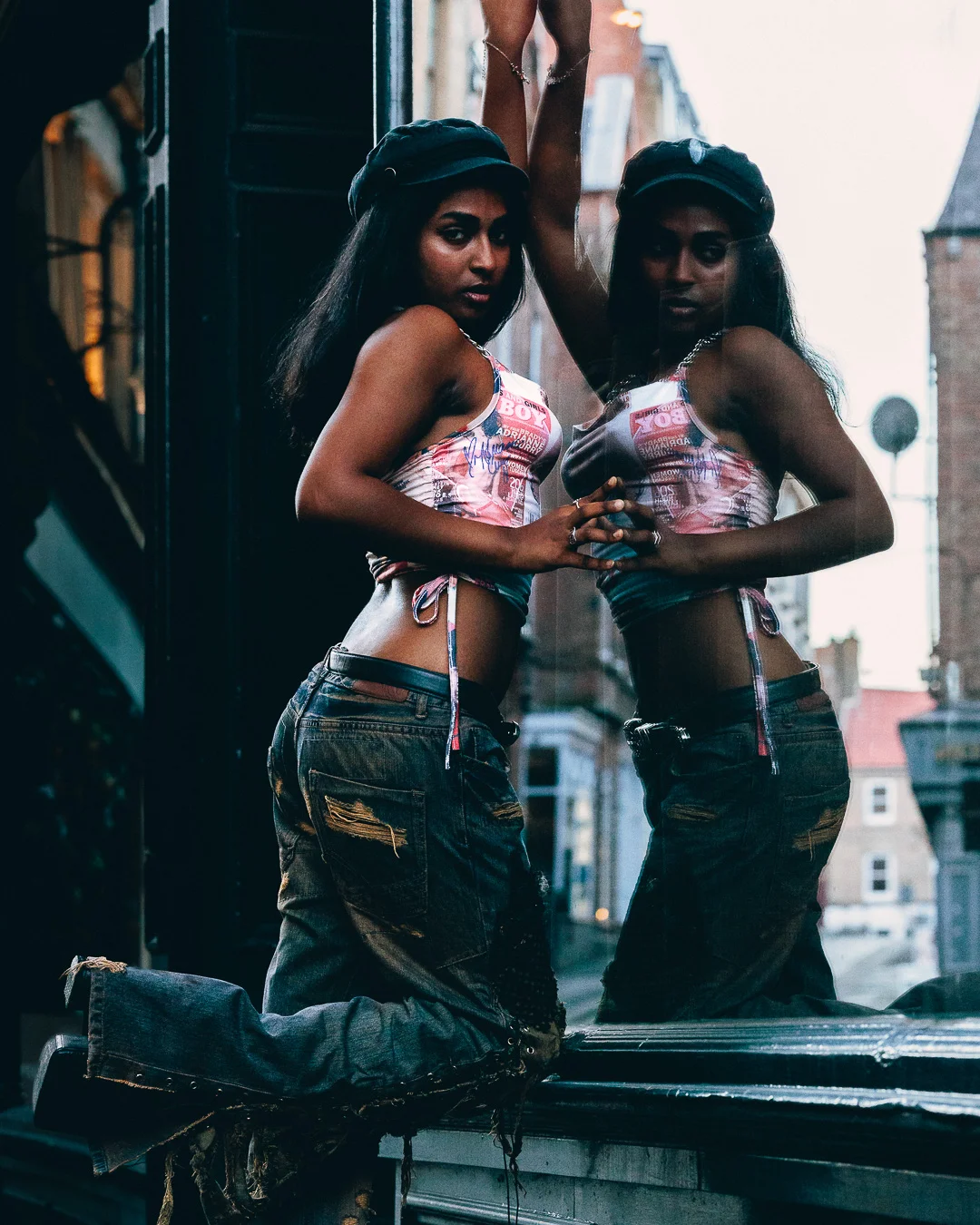 Woman in a crop top and distressed jeans posing with her reflection in an urban street setting.