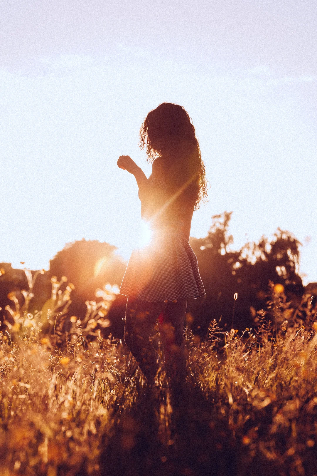 Silhouette of a woman with curly hair standing in a golden field at sunset, backlit by the sun.