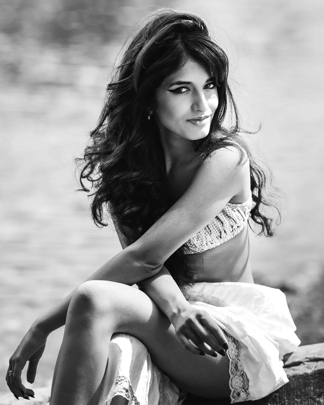 Black and white portrait of a woman with long wavy hair sitting outdoors in a bikini top and skirt.