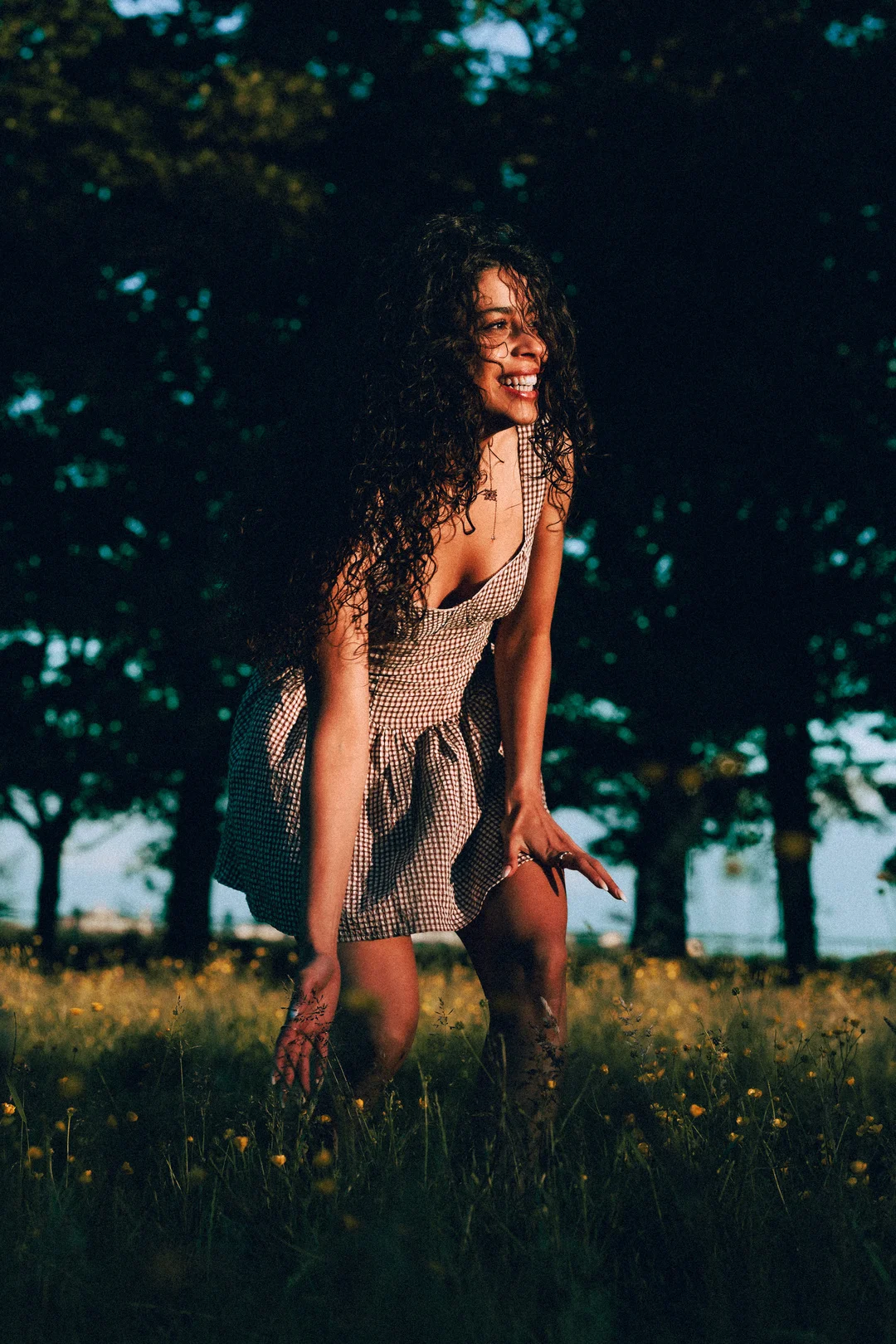 Smiling woman with curly hair in a checkered dress posing in a field of yellow flowers.