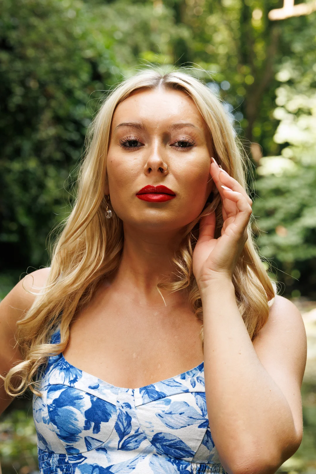 Close-up portrait of a blonde woman with red lipstick in a blue floral dress, posing against a green forest background.