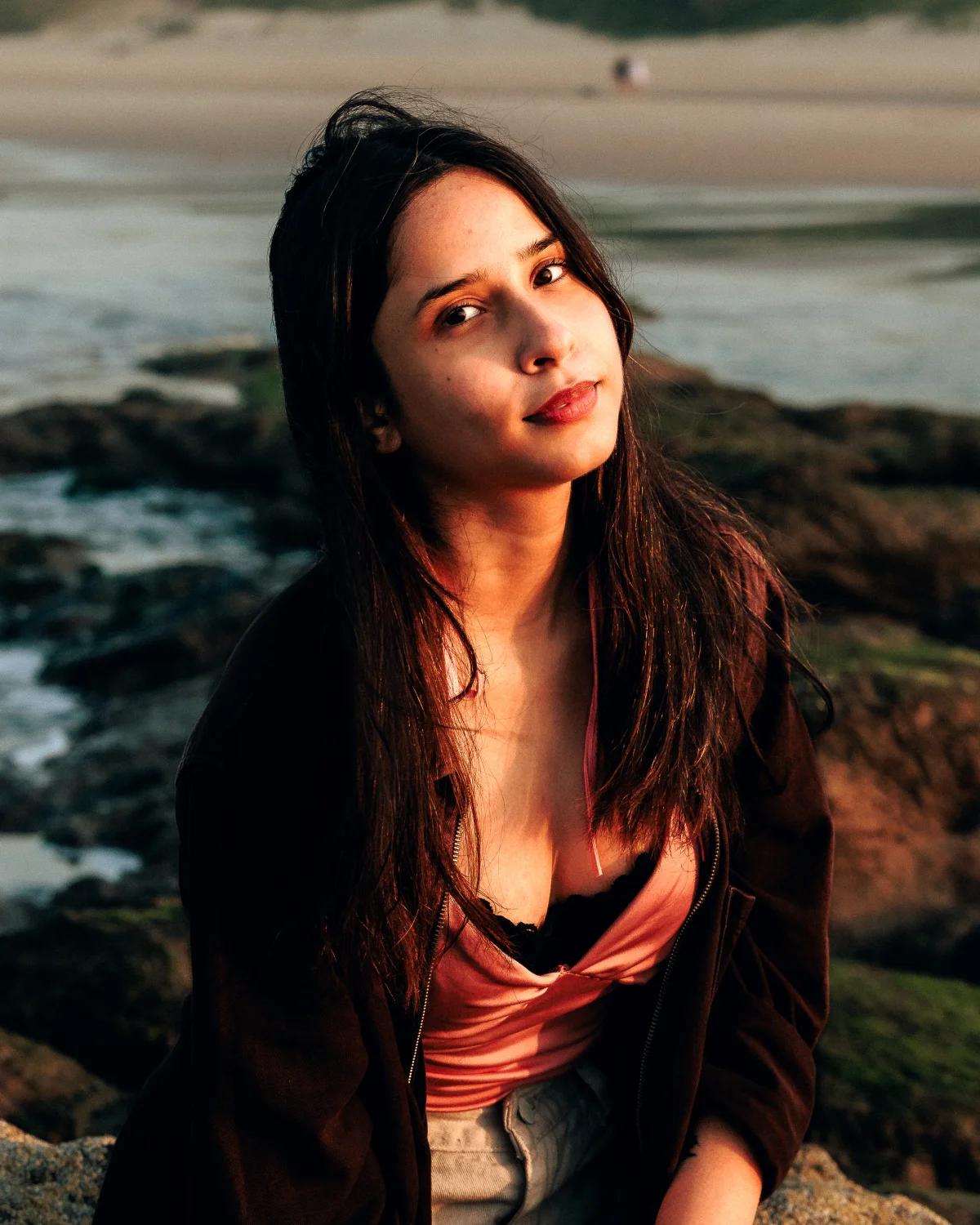 Portrait of a young woman with long dark hair posing on a rocky beach during golden hour.