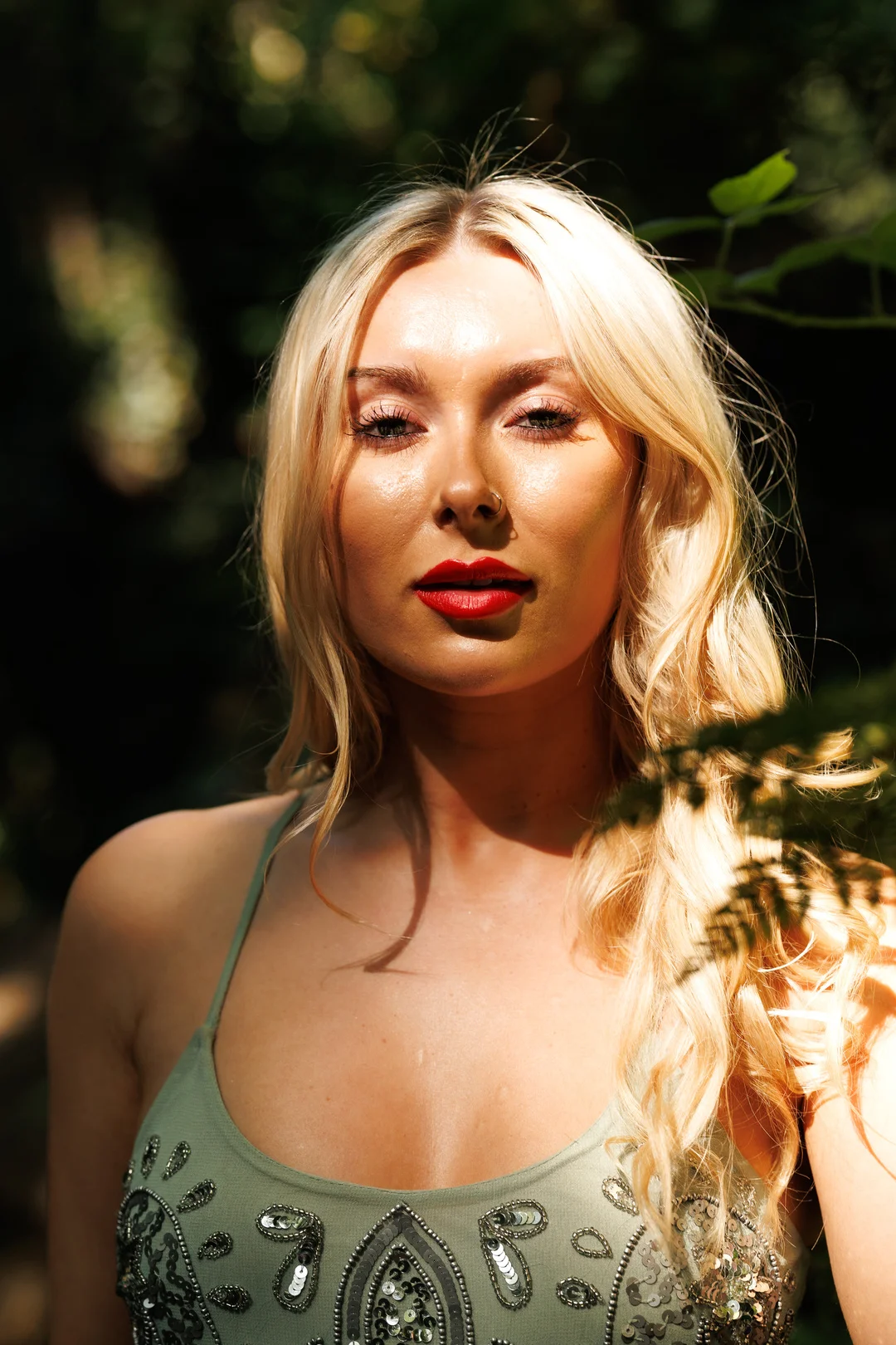 Close-up portrait of a blonde woman with red lipstick in a green sequined top, outdoors in natural sunlight.
