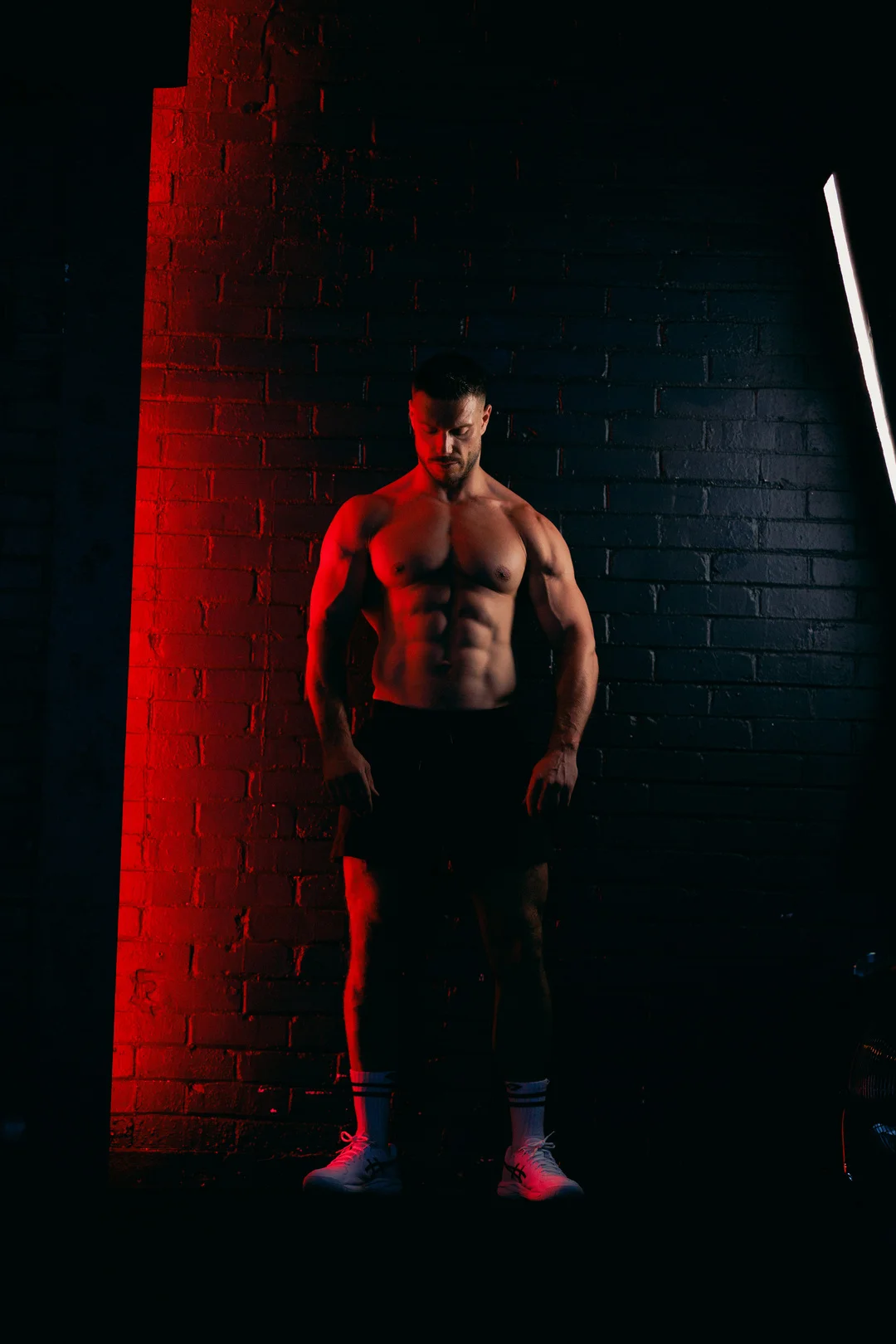 A muscular man posing shirtless against a dark brick wall with dramatic red and white lighting.