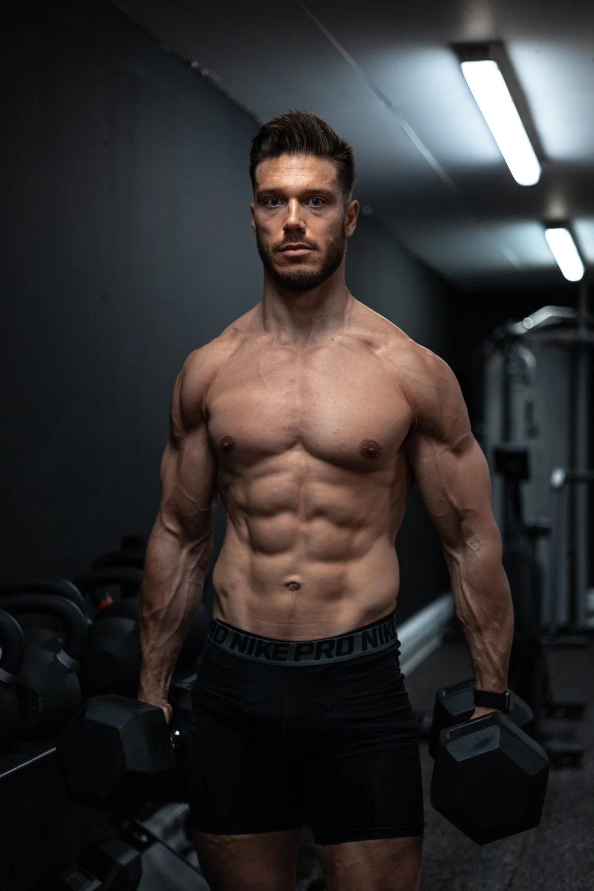 Muscular man holding dumbbells in a moody gym setting, shirtless and wearing compression shorts.