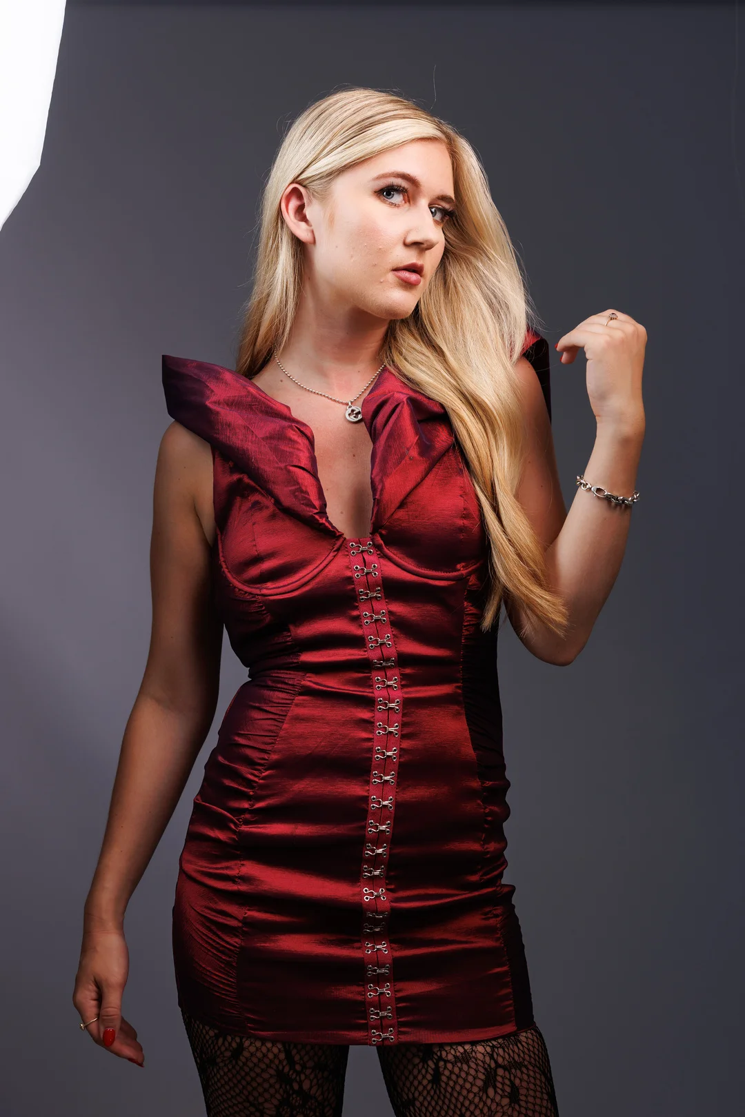 A blonde woman in a red satin dress and fishnets posing against a grey studio background.