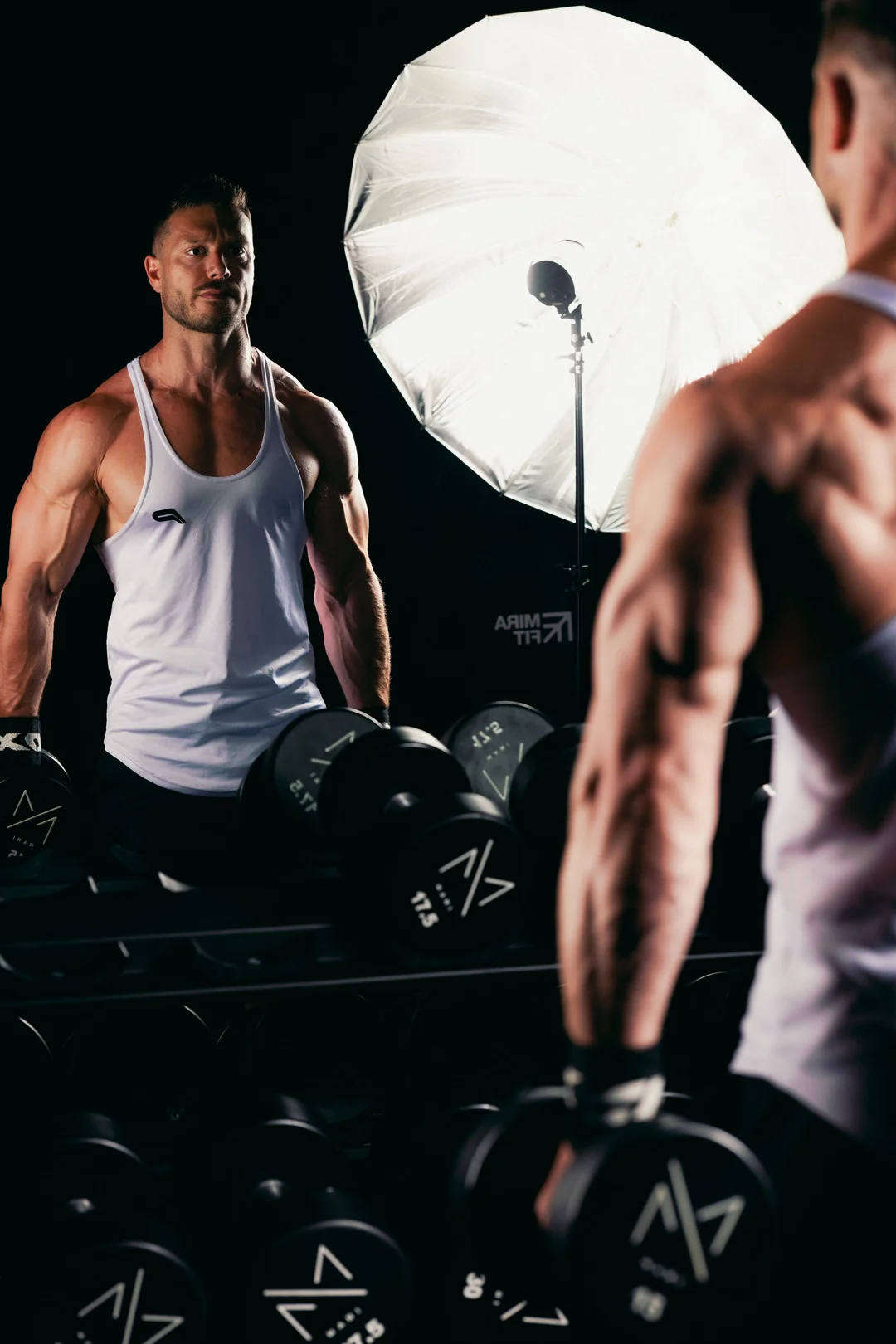 Muscular man in a white tank top posing in a gym, captured via a mirror reflection with studio lighting.