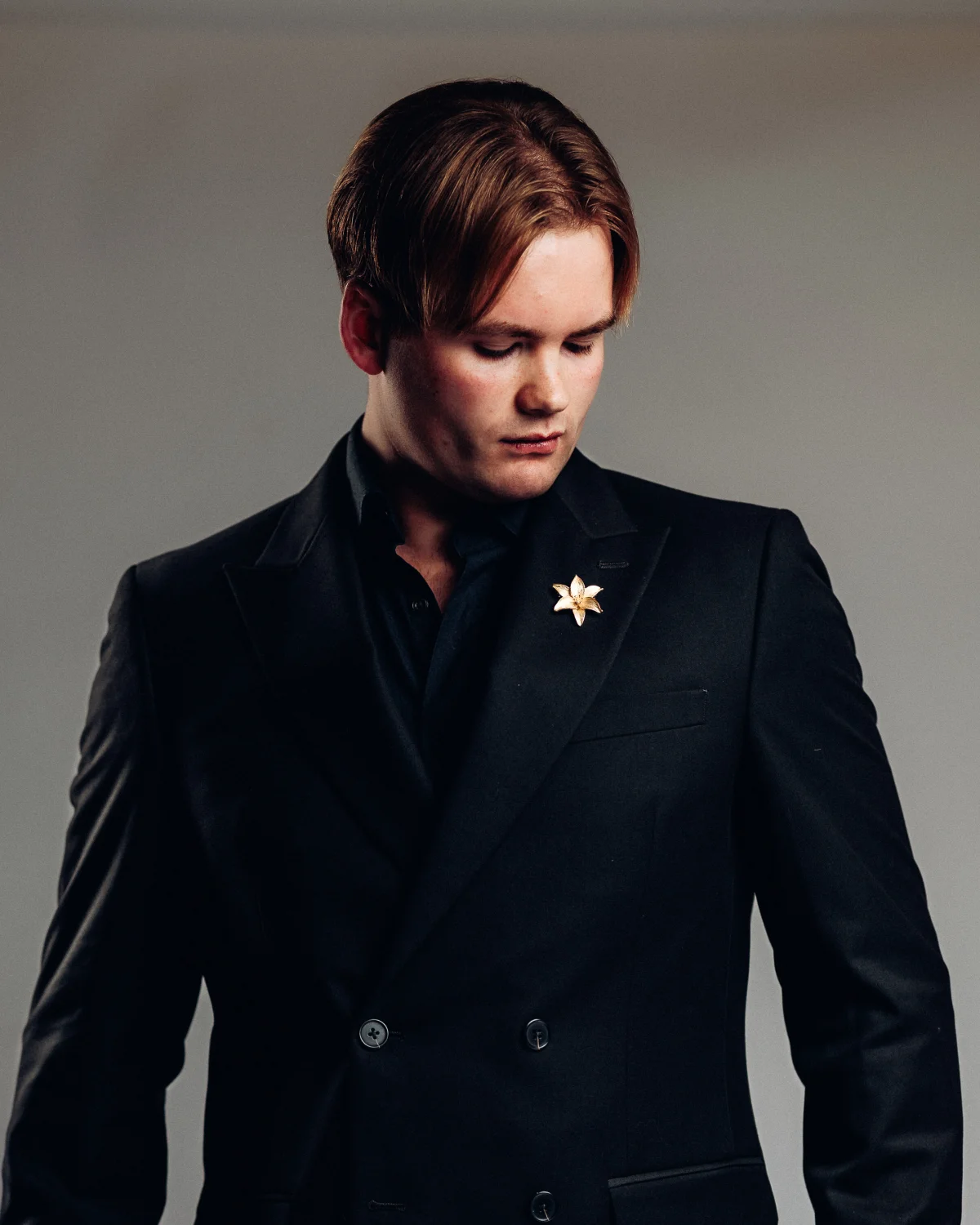 A man in a dark double-breasted suit with a gold flower pin, looking down against a neutral background.