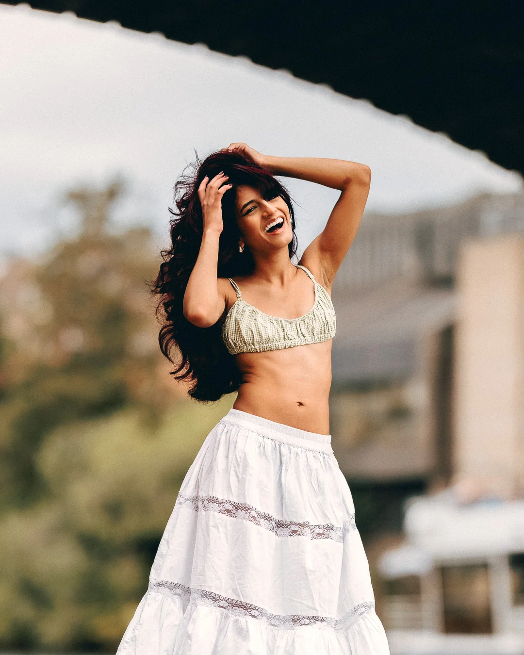 A joyful woman with long dark hair wearing a light crop top and white tiered skirt, posing outdoors.