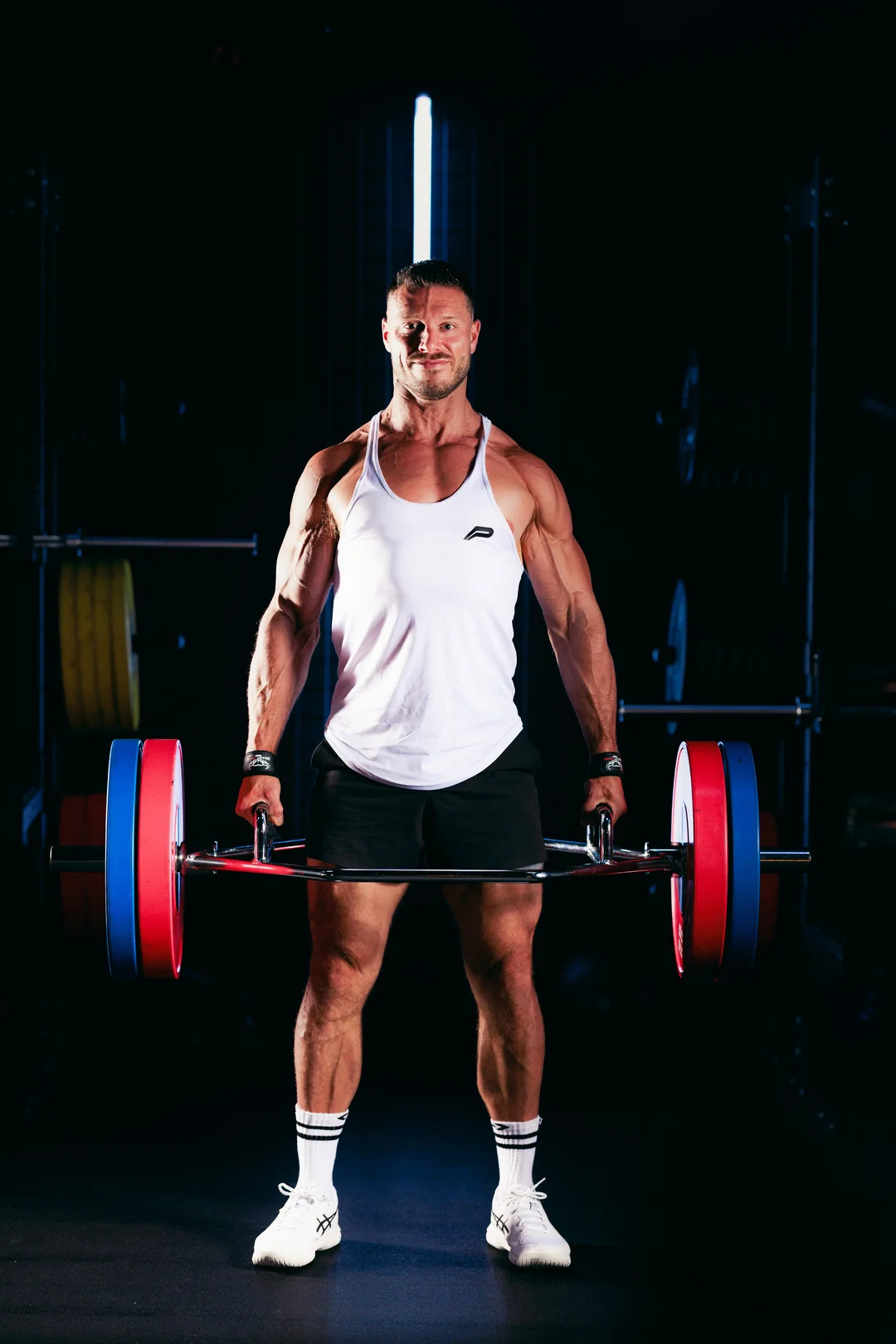 Muscular man in a white tank top holding a barbell in a gym with dramatic, high-contrast lighting.