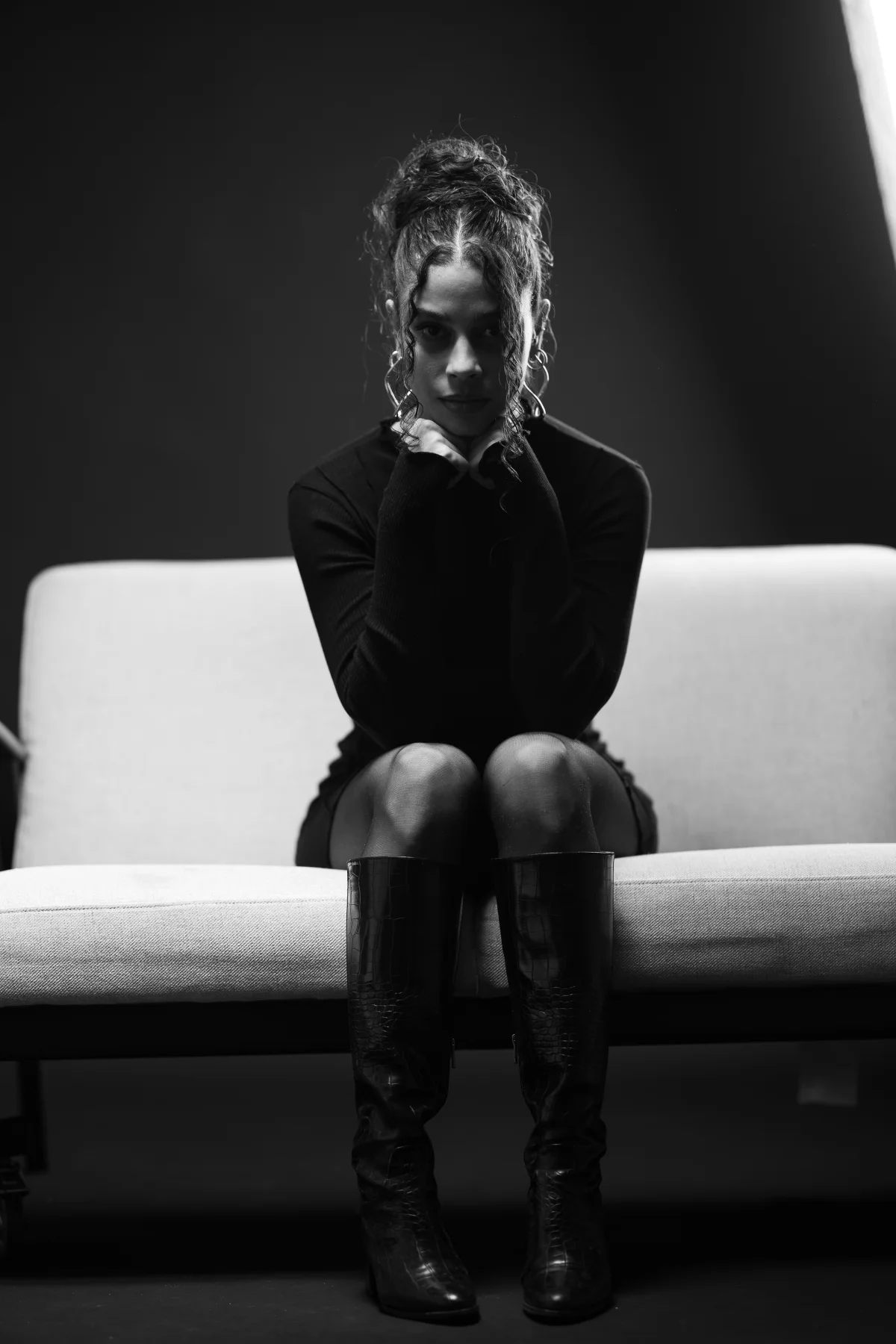 Black and white fashion portrait of a woman in a black outfit and tall boots sitting on a white couch.
