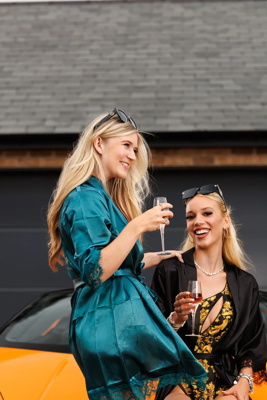 Two women in satin robes holding champagne glasses, posing in front of an orange luxury car.