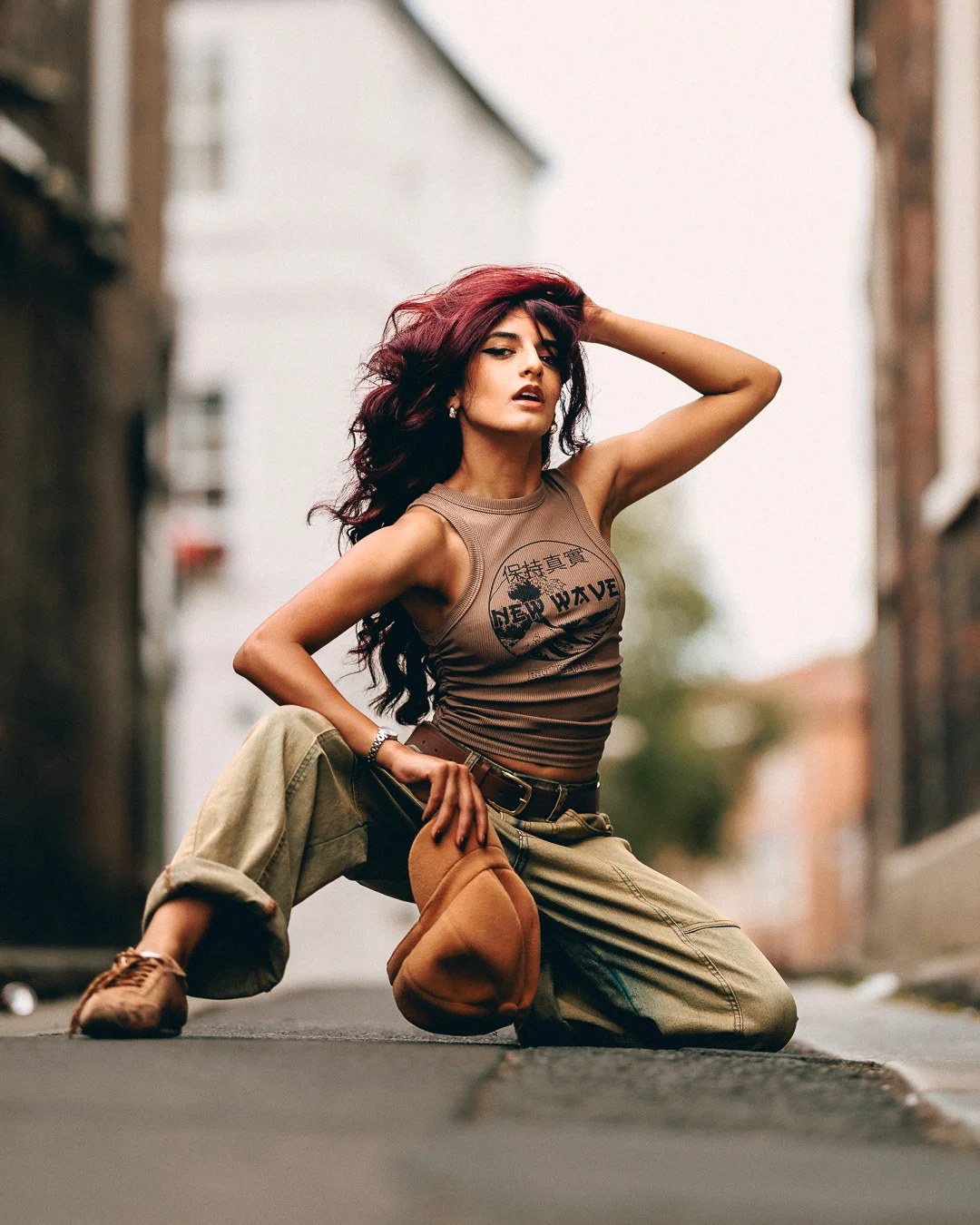 A woman with vibrant red hair posing in an urban alley, wearing a tan tank top and olive cargo pants.