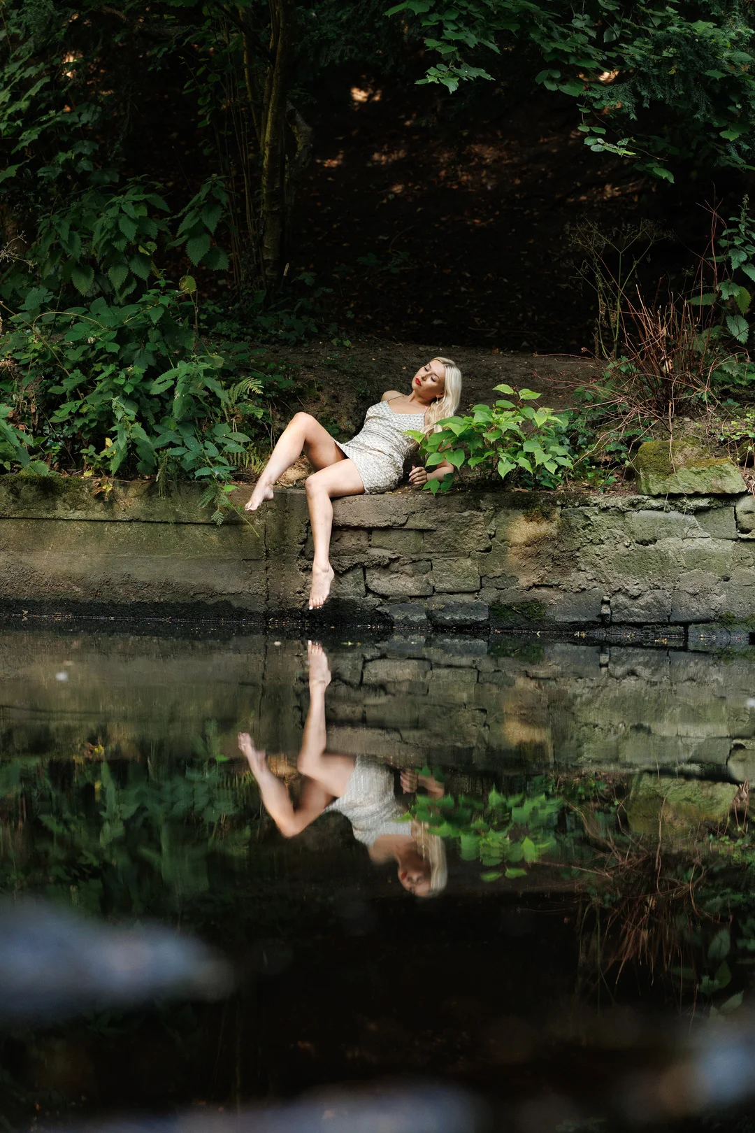 A blonde woman in a short white dress reclining on a stone wall by a pond, reflecting in the water.