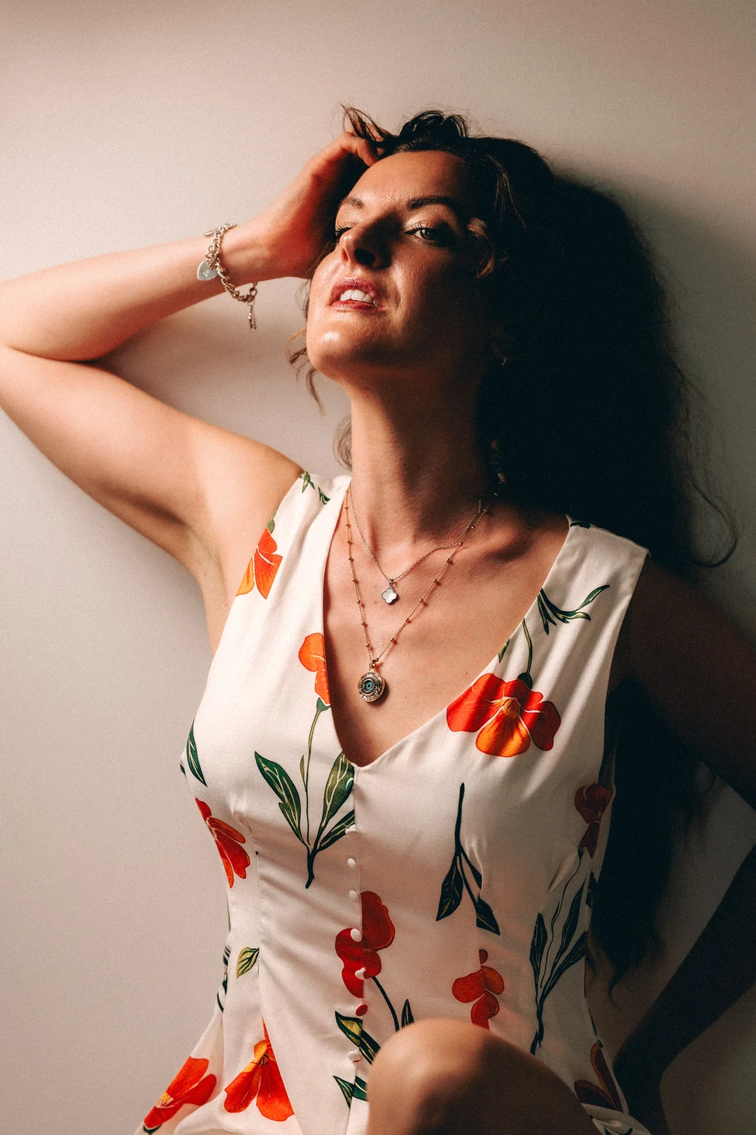 A woman in a white floral dress posing against a light wall with her hand in her hair.