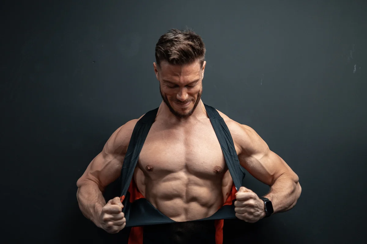 Muscular man pulling up his black tank top to reveal defined abs, smiling against a dark grey background.