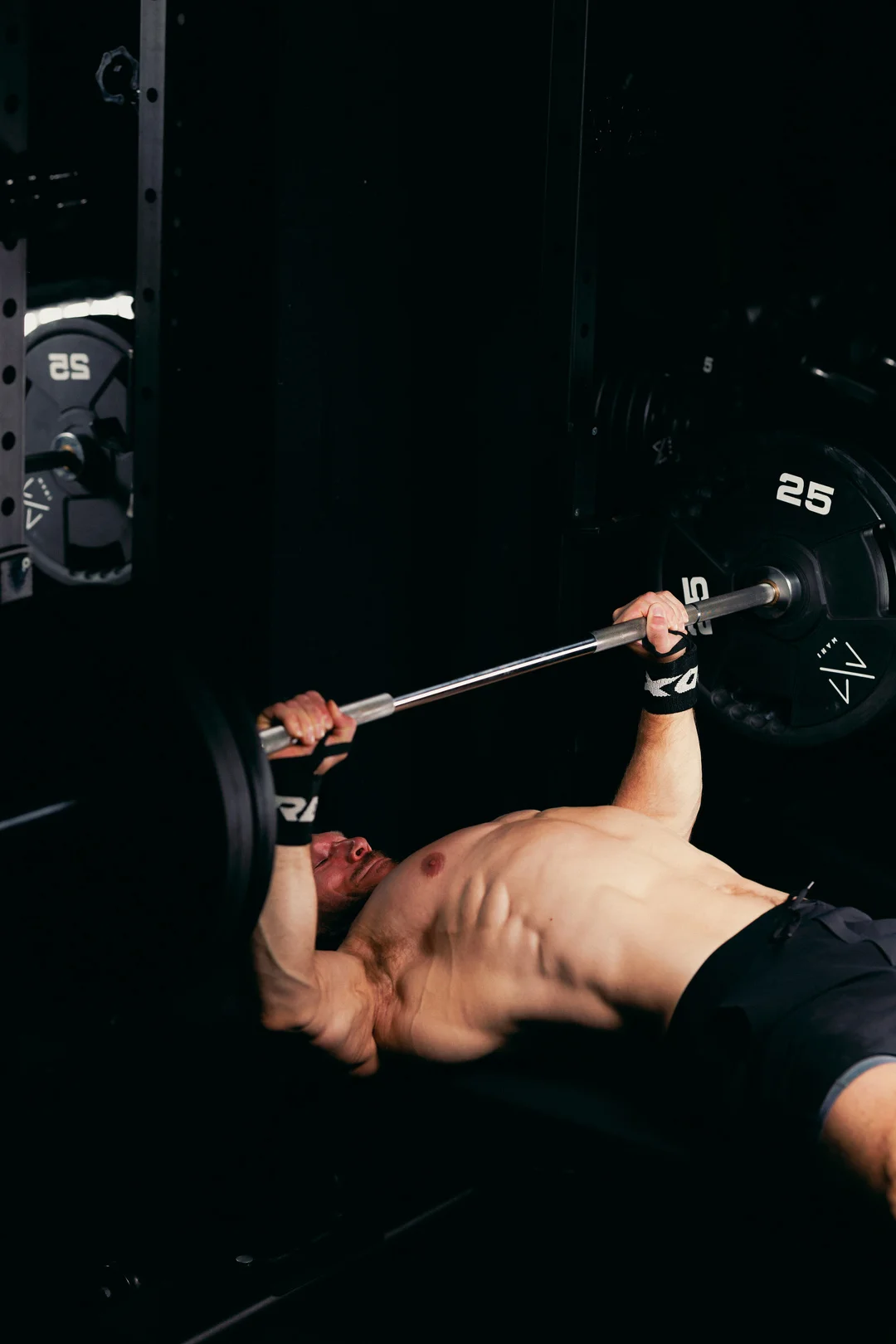 A shirtless man performing a bench press in a dark gym, capturing a moment of intense physical effort.