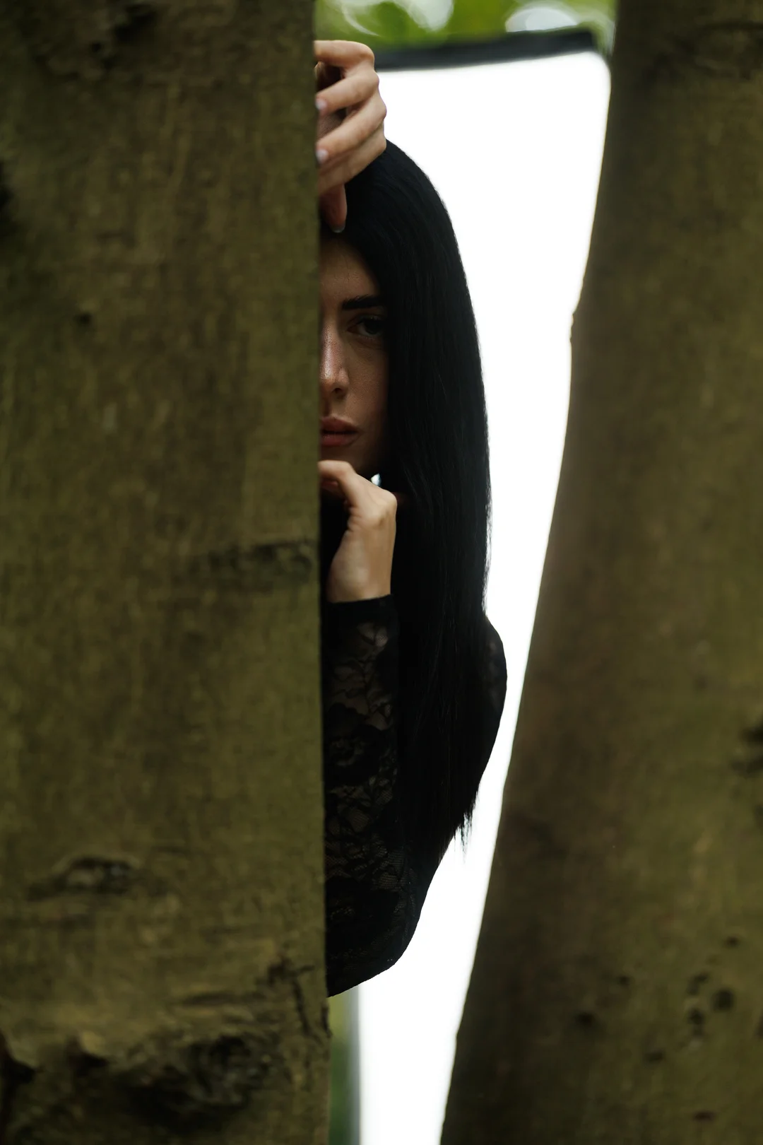 A woman with long black hair peeking from behind a tree trunk in a moody outdoor setting.
