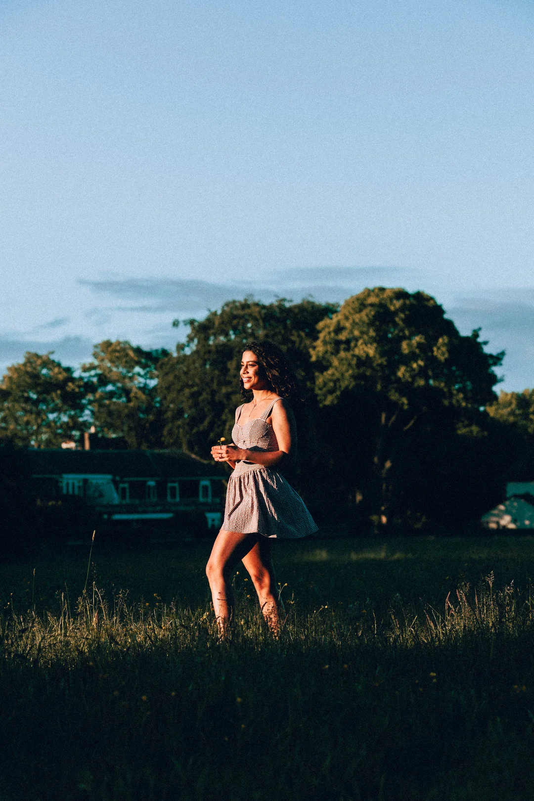 A smiling woman in a red and white checkered dress standing in a grassy field at golden hour.