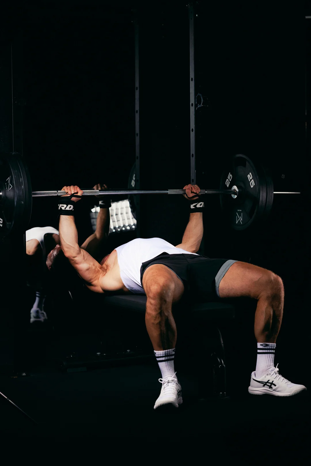 A muscular man performing a bench press in a dark gym, with a mirror reflection.