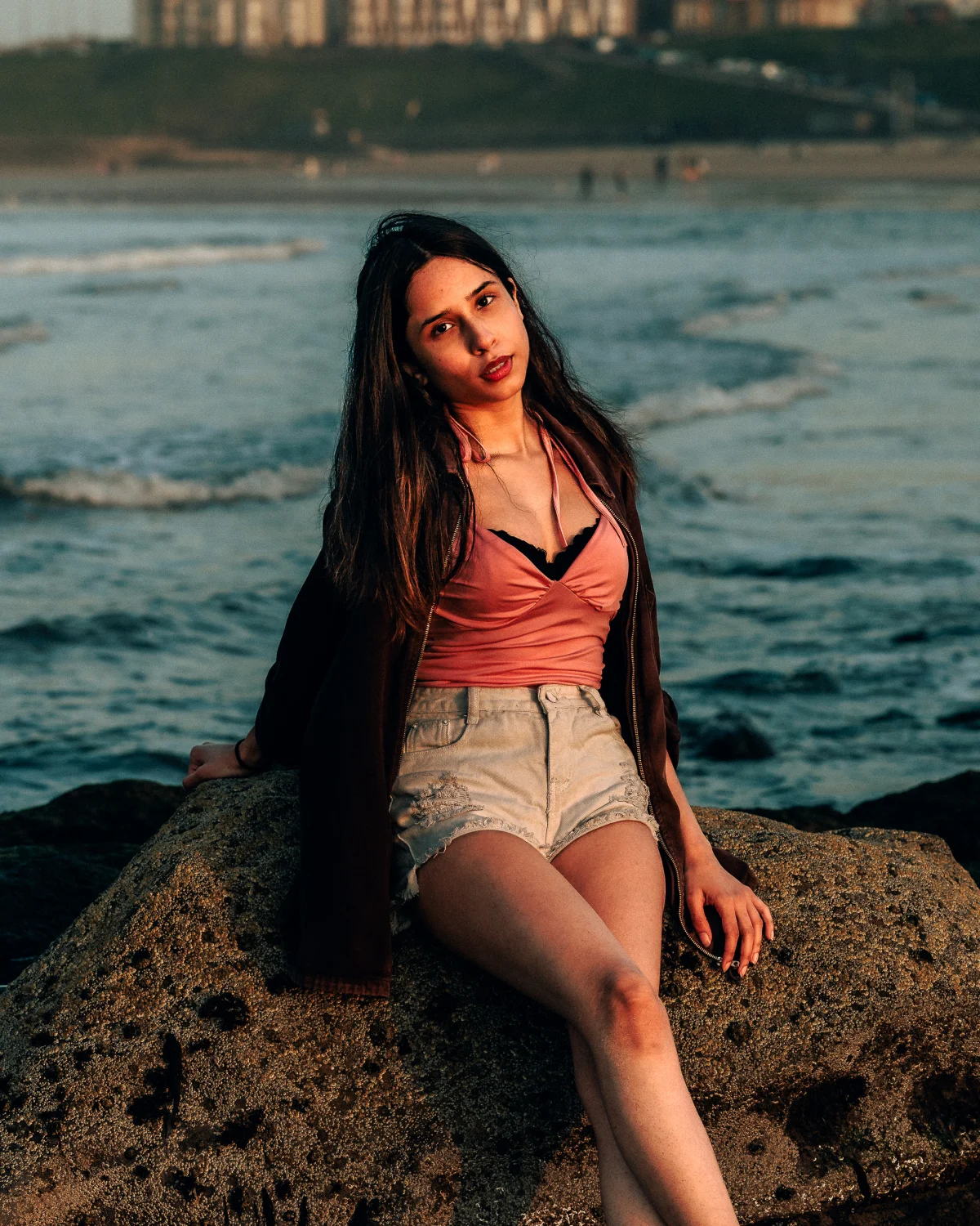 Young woman with long dark hair posing on a rock at a beach during sunset, wearing a pink top and denim shorts.