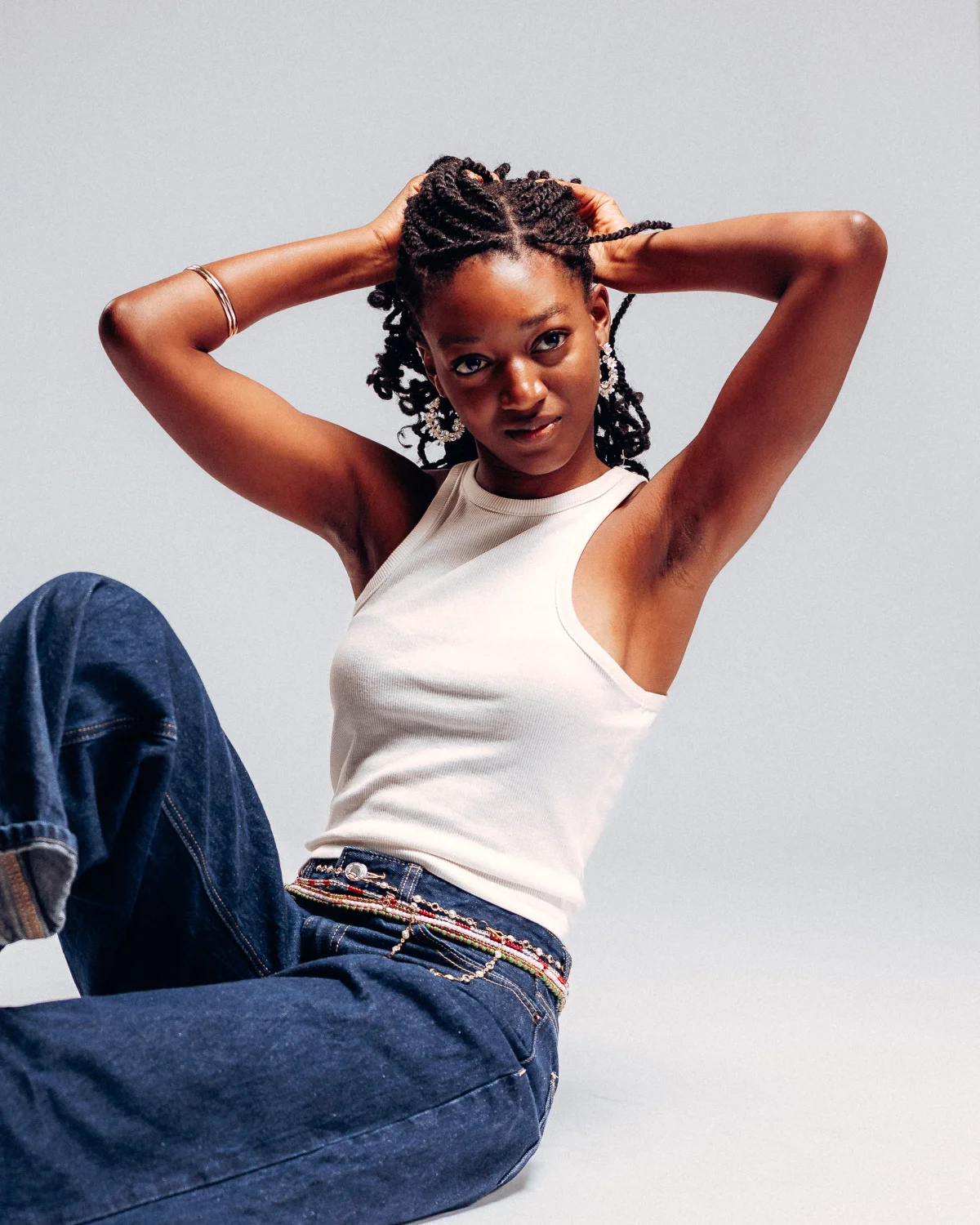A Black woman with braided hair posing in a white tank top and blue jeans against a light grey studio background.