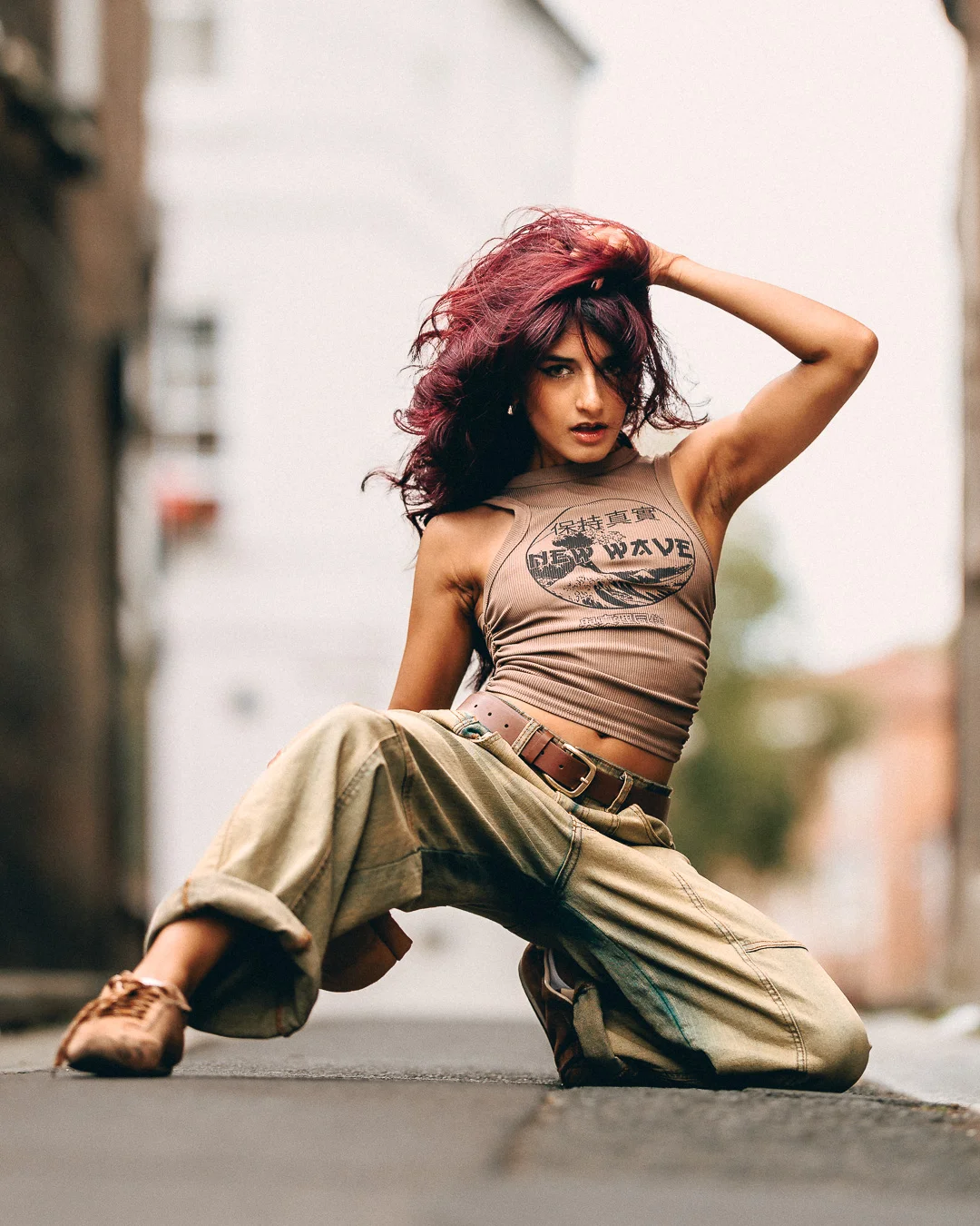 Woman with red hair in a tan crop top and cargo pants posing in an urban alleyway.